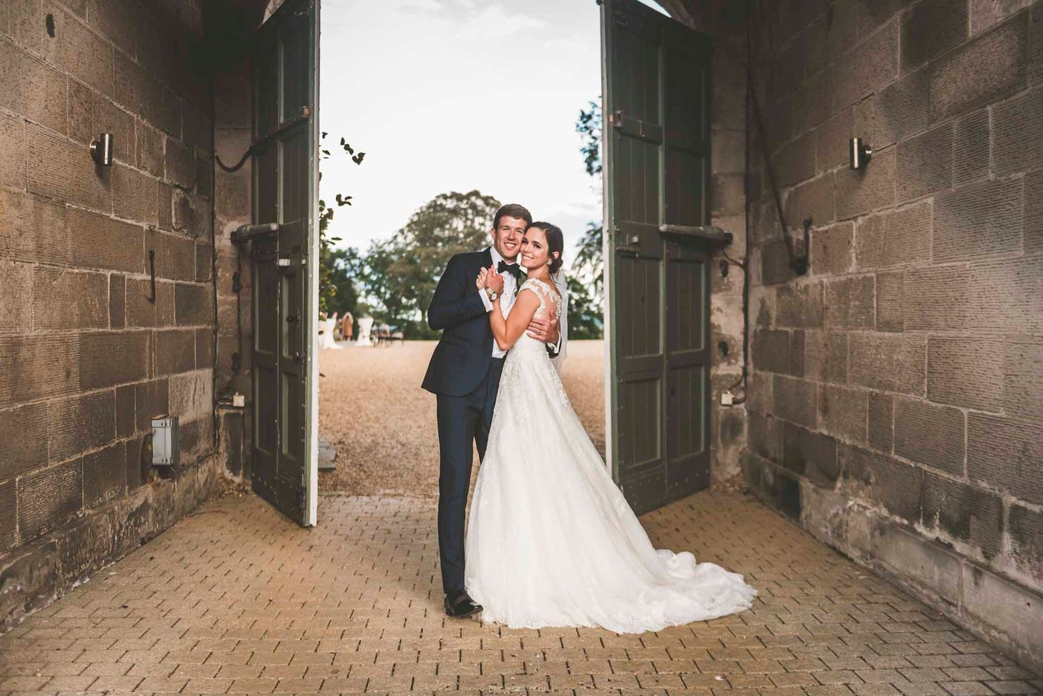 Bride and groom embracing in a rustic stone doorway, bride in a flowing white gown with train, groom in a dark suit.