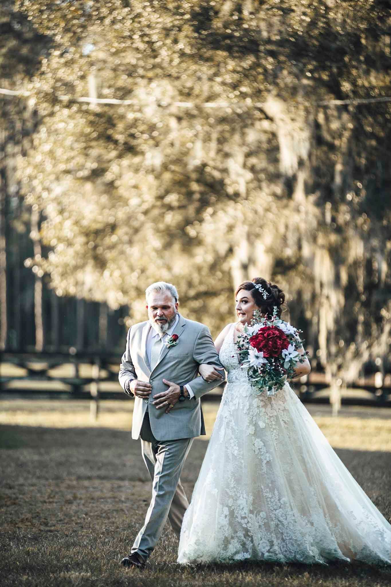 Bride in lace gown holding red roses walks arm-in-arm with man in gray suit outdoors among golden trees.