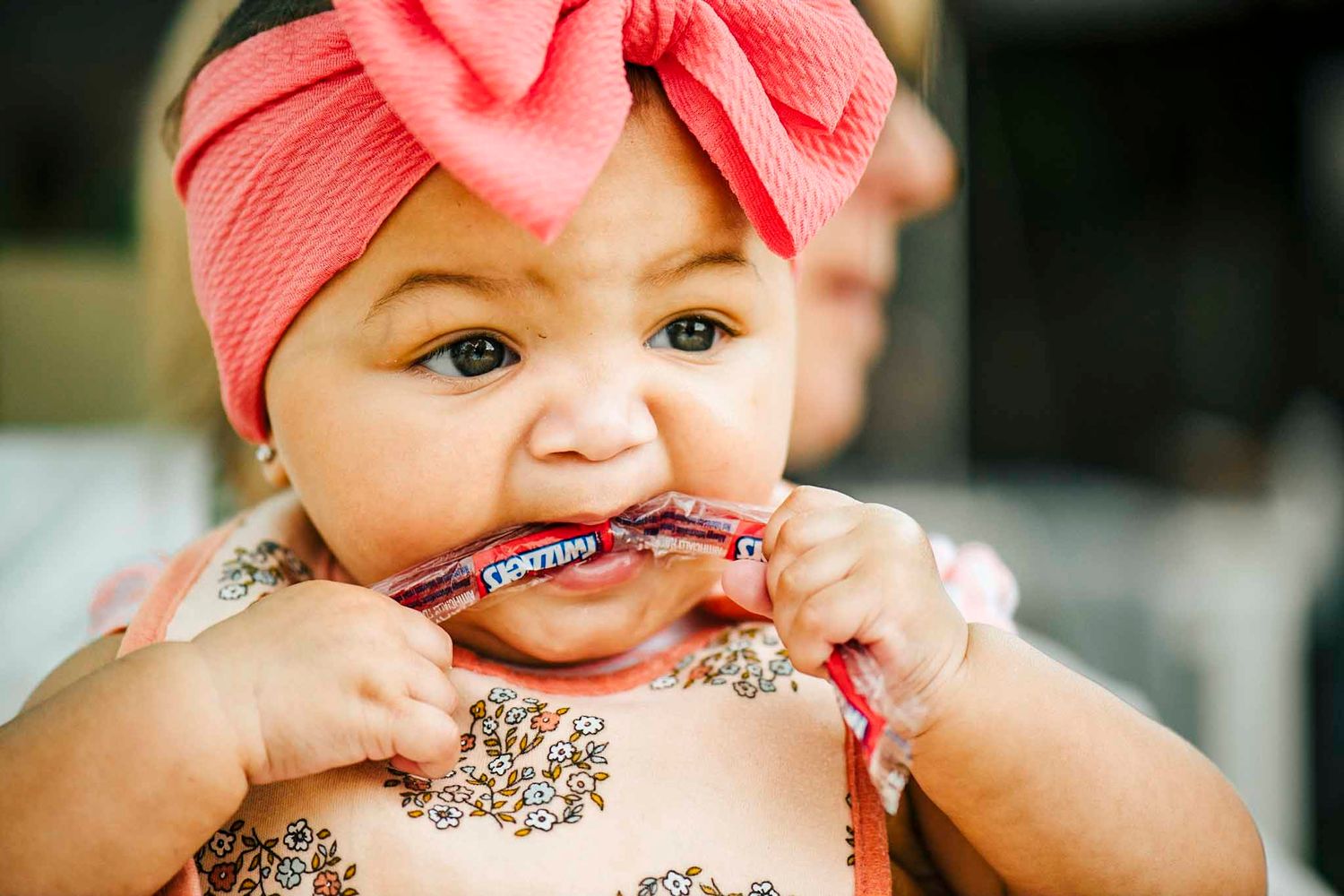 Adorable baby girl in pink bow headband and sparkly outfit chewing on a Cracker Jack box.