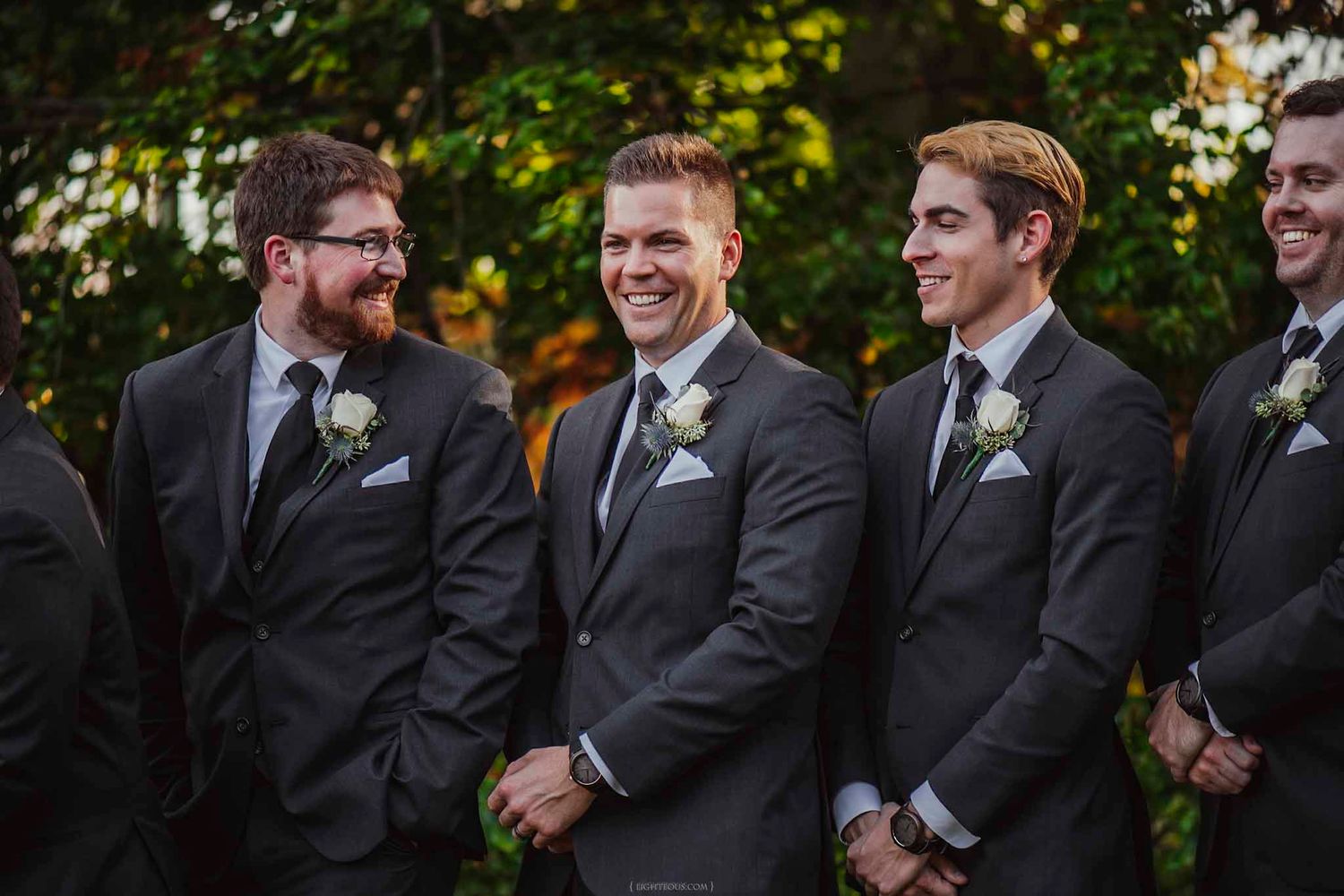 Groom and groomsmen in gray suits with white boutonnieres laughing together outdoors at a wedding.