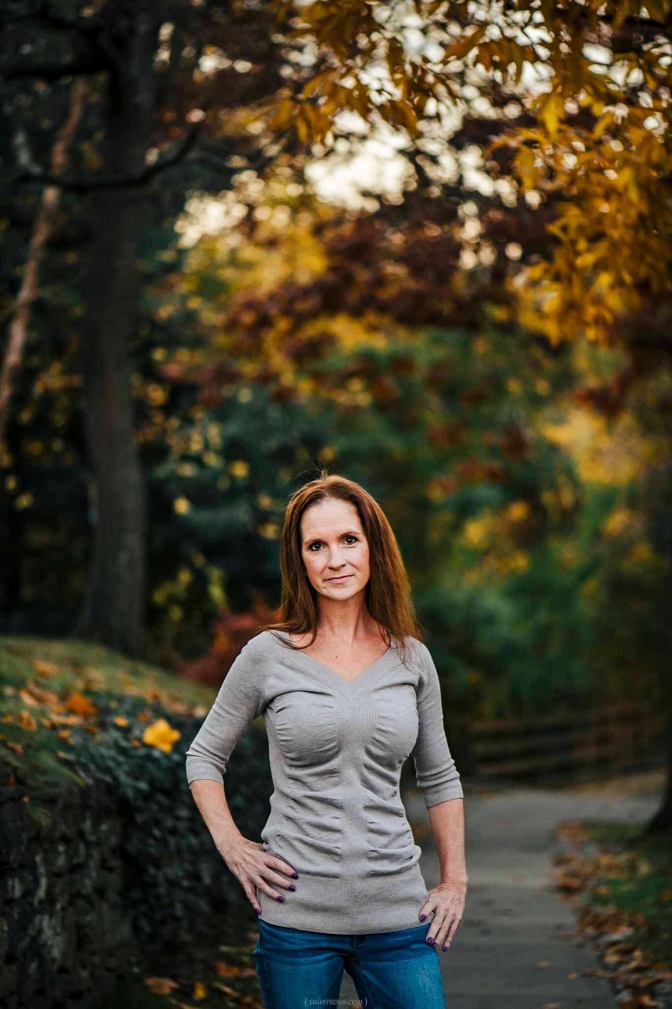 A confident woman in a gray top and jeans poses outdoors among vibrant autumn foliage on a scenic park path.