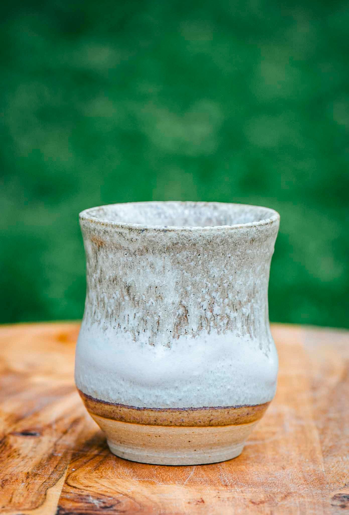 Handcrafted stoneware ceramic cup with white speckled glaze on wooden surface, green background.