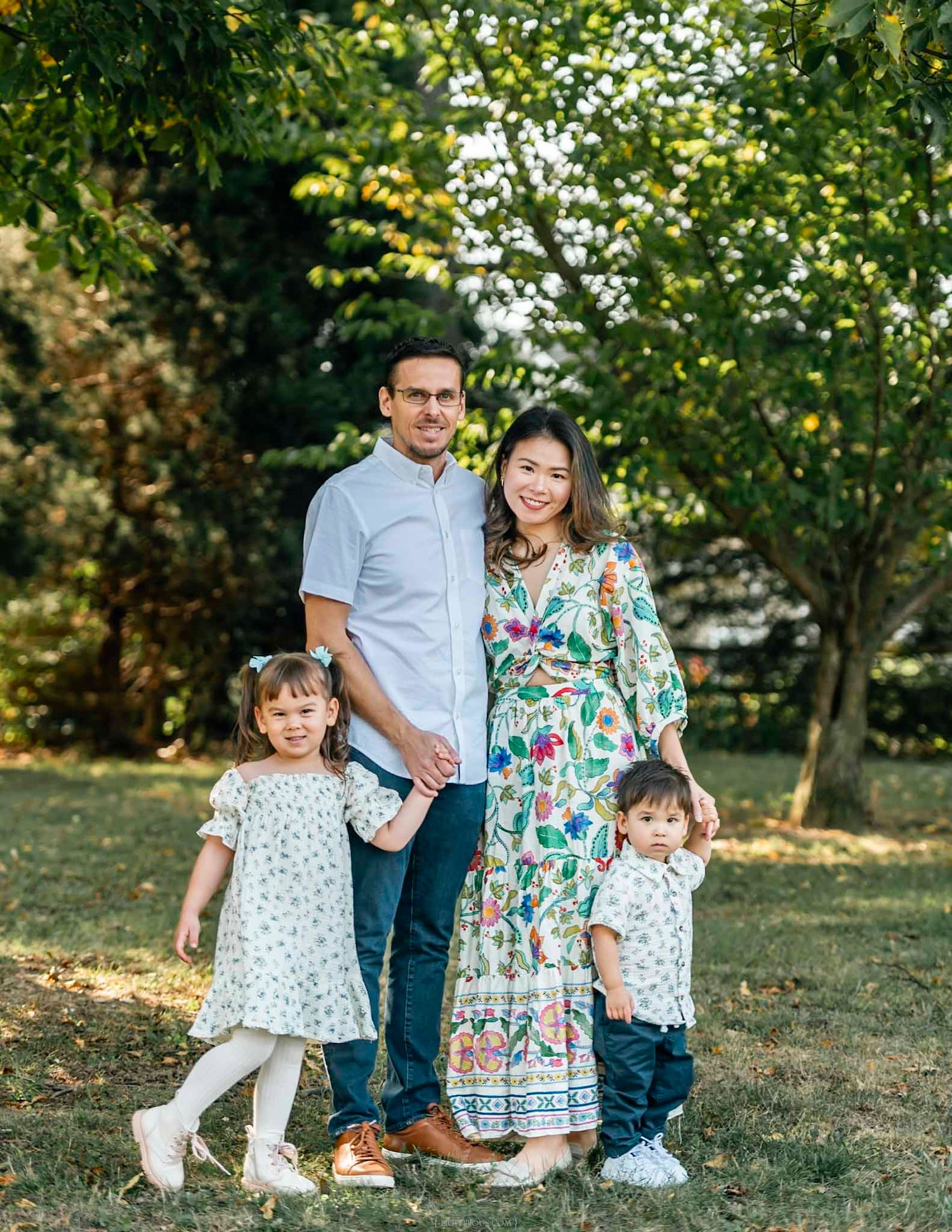 A smiling family of four poses outdoors in a lush green park, with two young children in floral outfits.