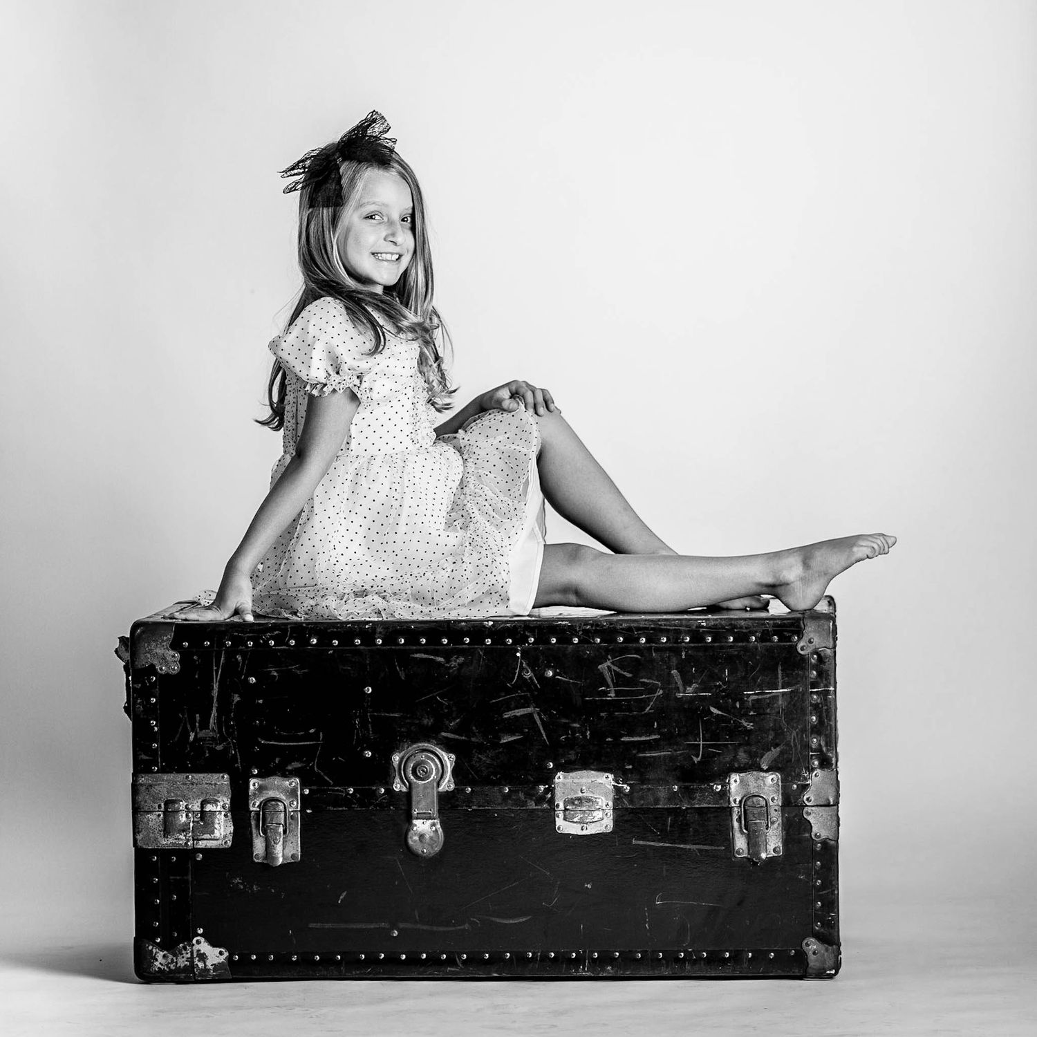 Black and white photo of a smiling young girl in a dress sitting on a vintage trunk.