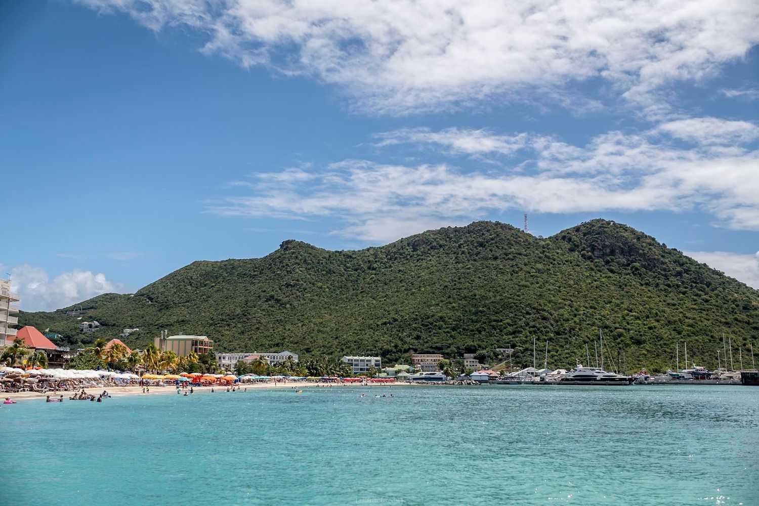 Turquoise Caribbean waters with a sandy beach, lush green mountain, and marina under a bright blue sky in St. Maarten.
