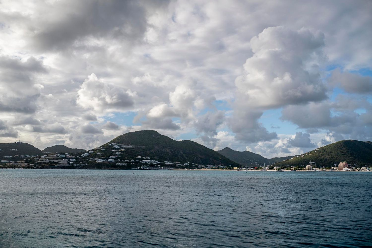 Scenic Caribbean island coastline with lush green mountains and cloudy sky reflecting on calm blue ocean waters.