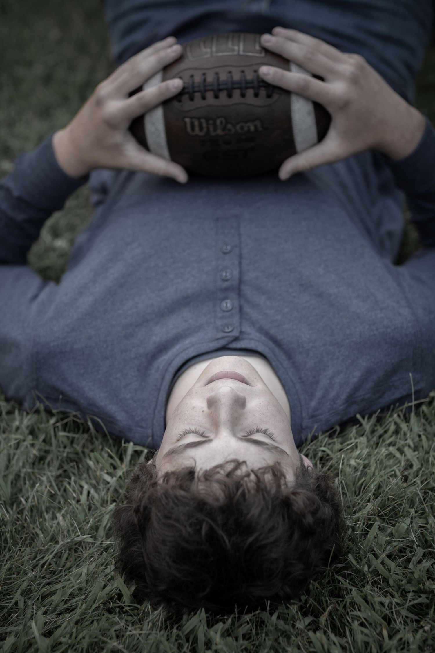 Teen boy lying on grass holding a Wilson football in a moody senior portrait photo.