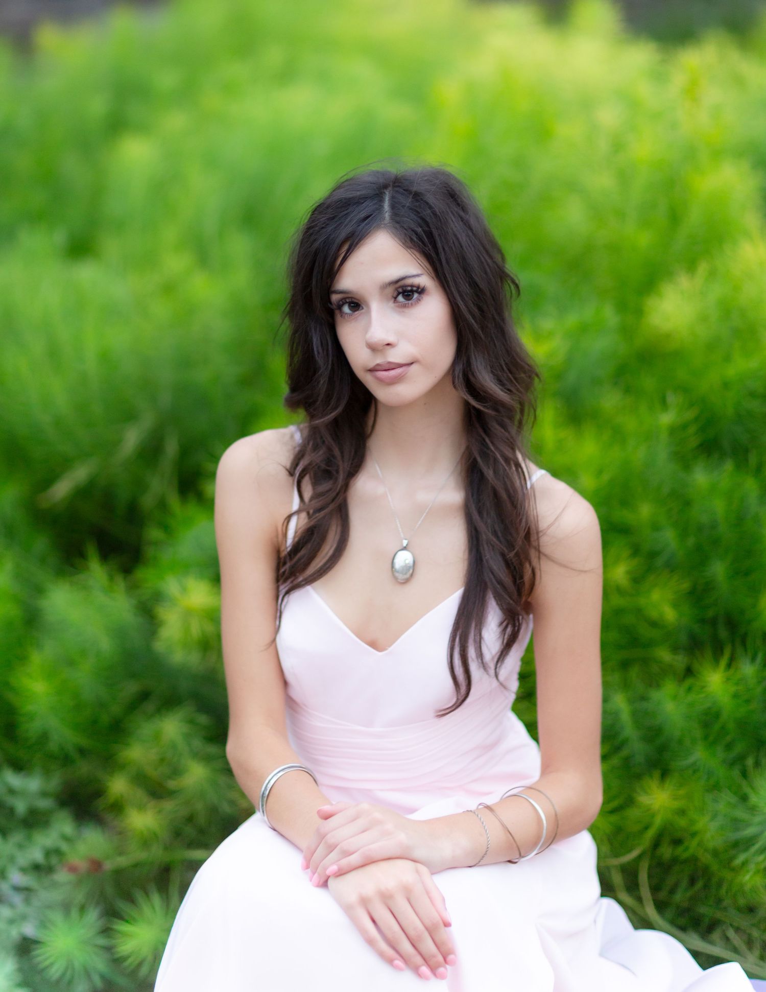 Young woman in pink dress with silver necklace and bracelet sitting outdoors surrounded by lush green foliage.