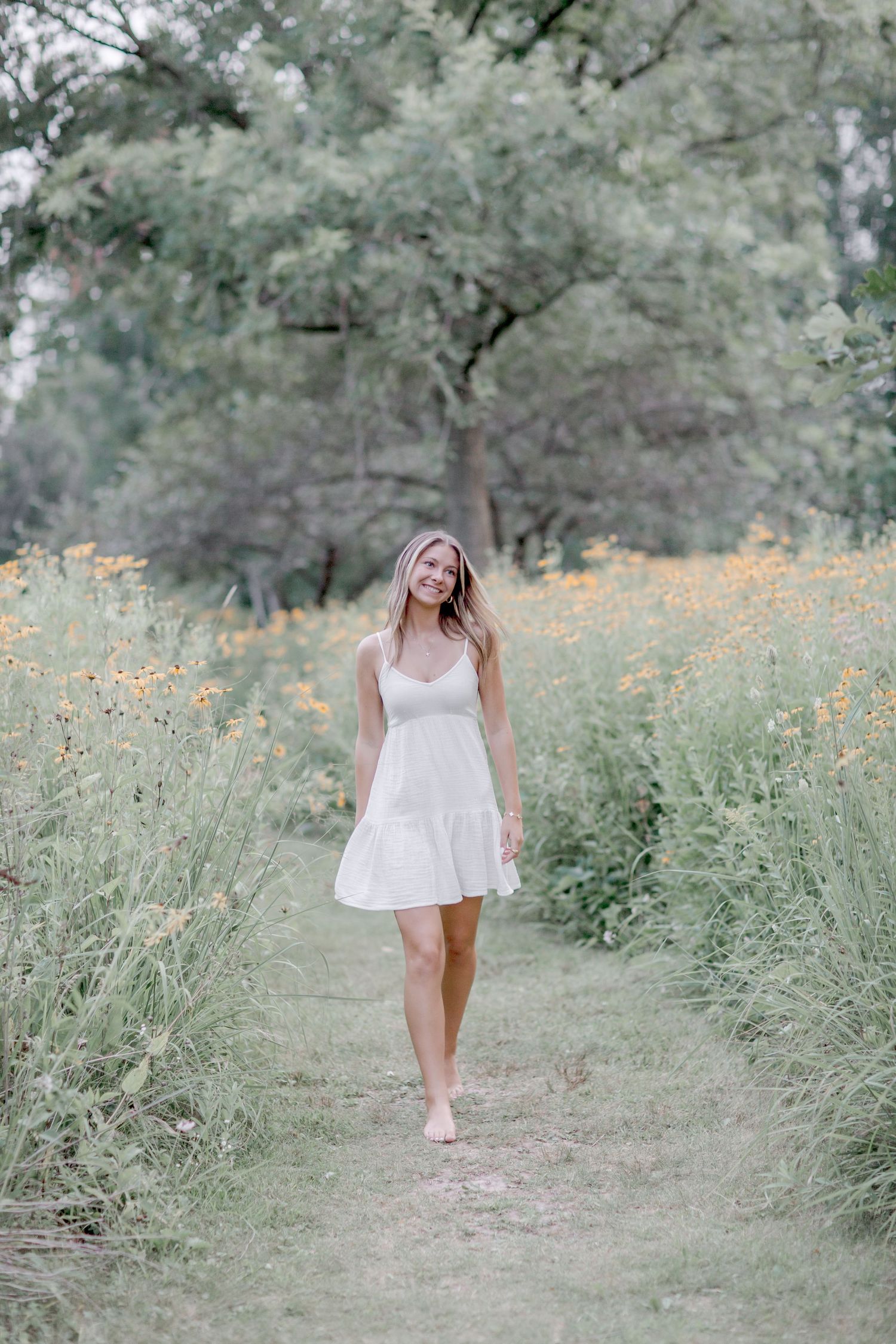 Smiling blonde woman in white sundress walking barefoot on a wildflower meadow path surrounded by yellow flowers.