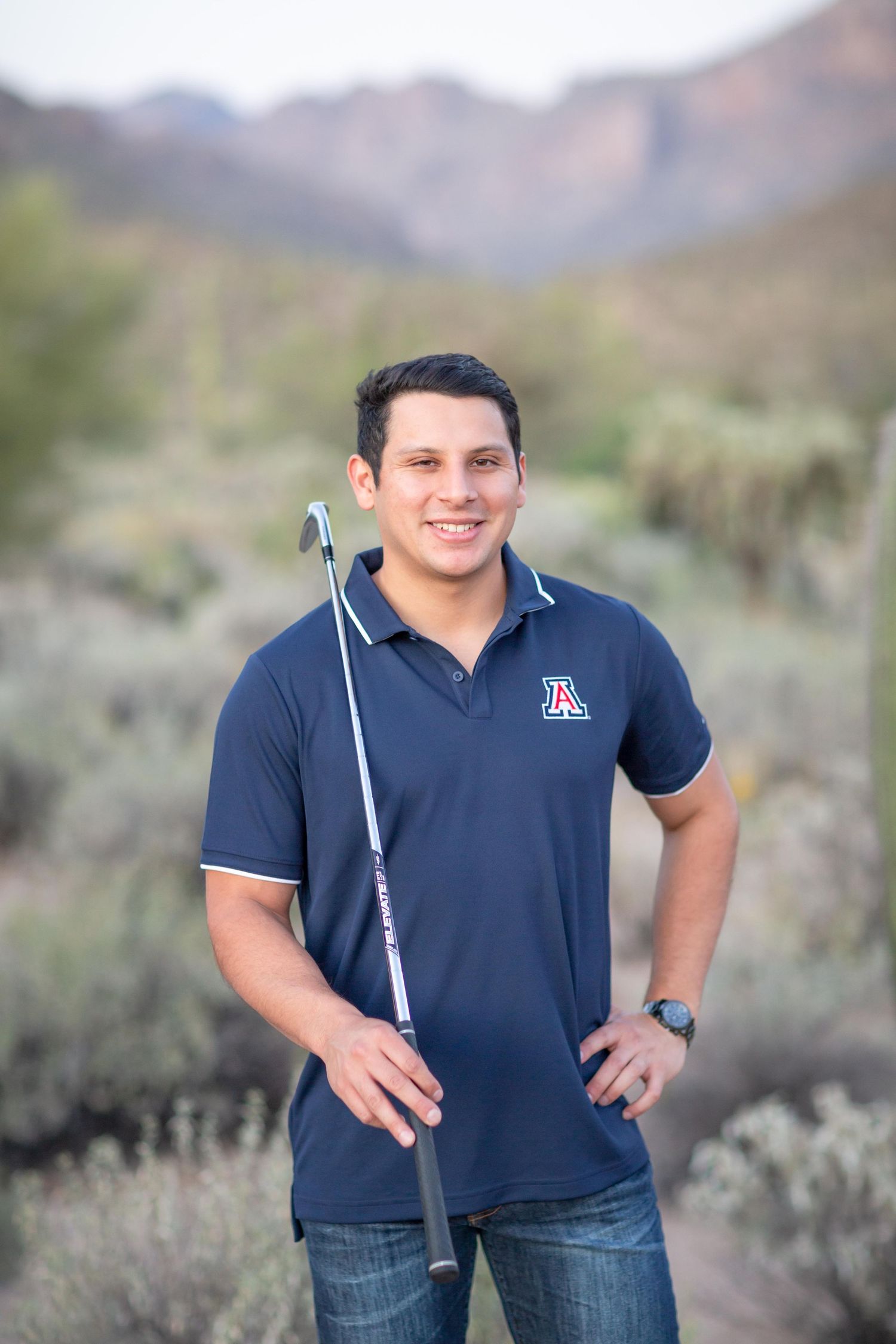 Young man in University of Arizona polo holding a golf club in desert landscape.