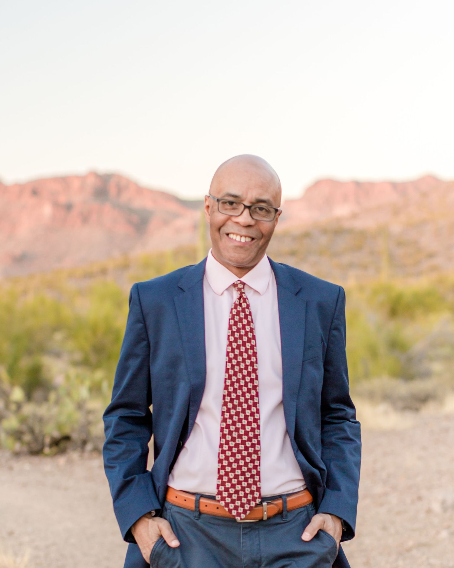Smiling bald man in navy suit and red tie standing in desert landscape at sunset.
