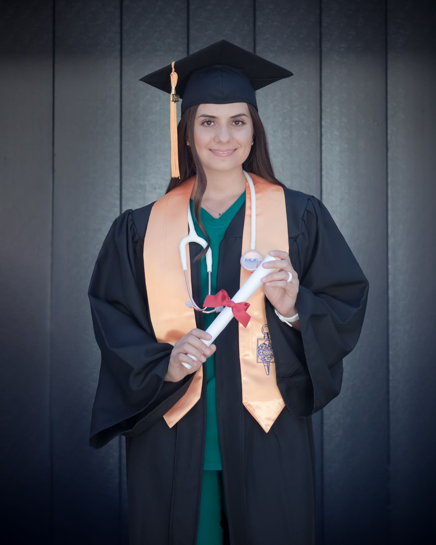 Female graduate in black cap and gown holding diploma with orange honor stole and stethoscope.