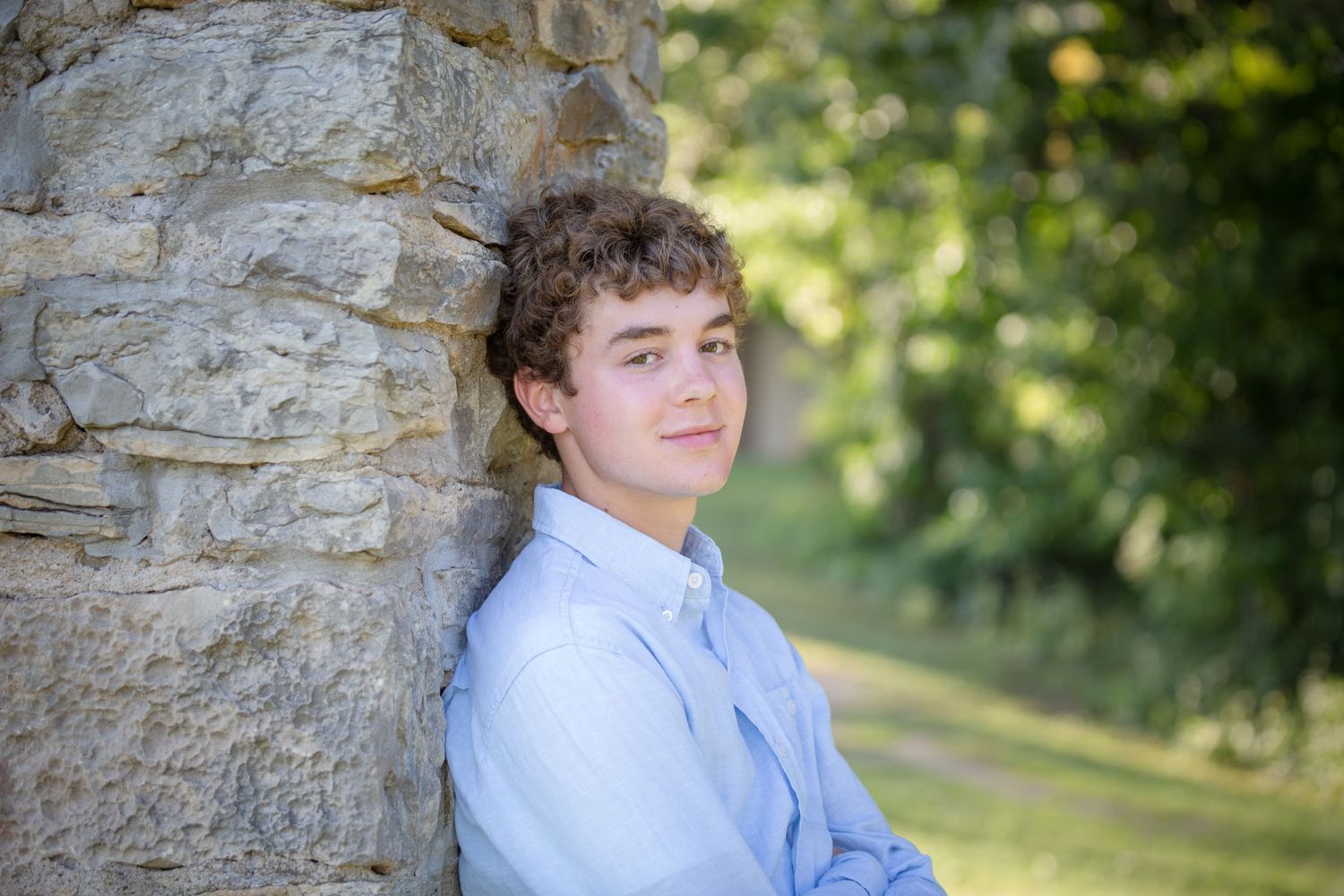 A teenage boy with curly hair leans against a stone wall outdoors, smiling with arms crossed in a blue shirt.