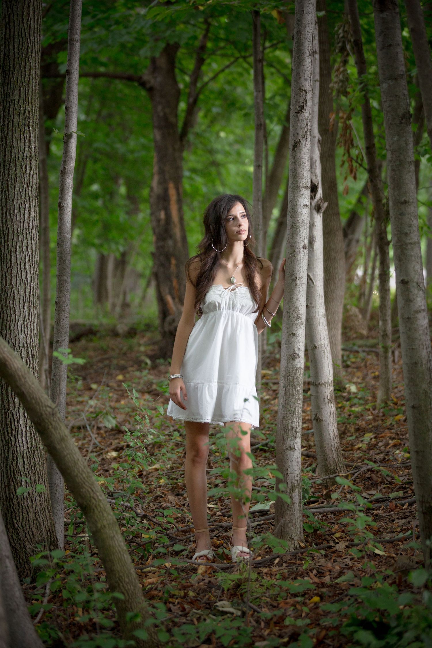 Young woman in white sundress standing among tall trees in a lush green forest.