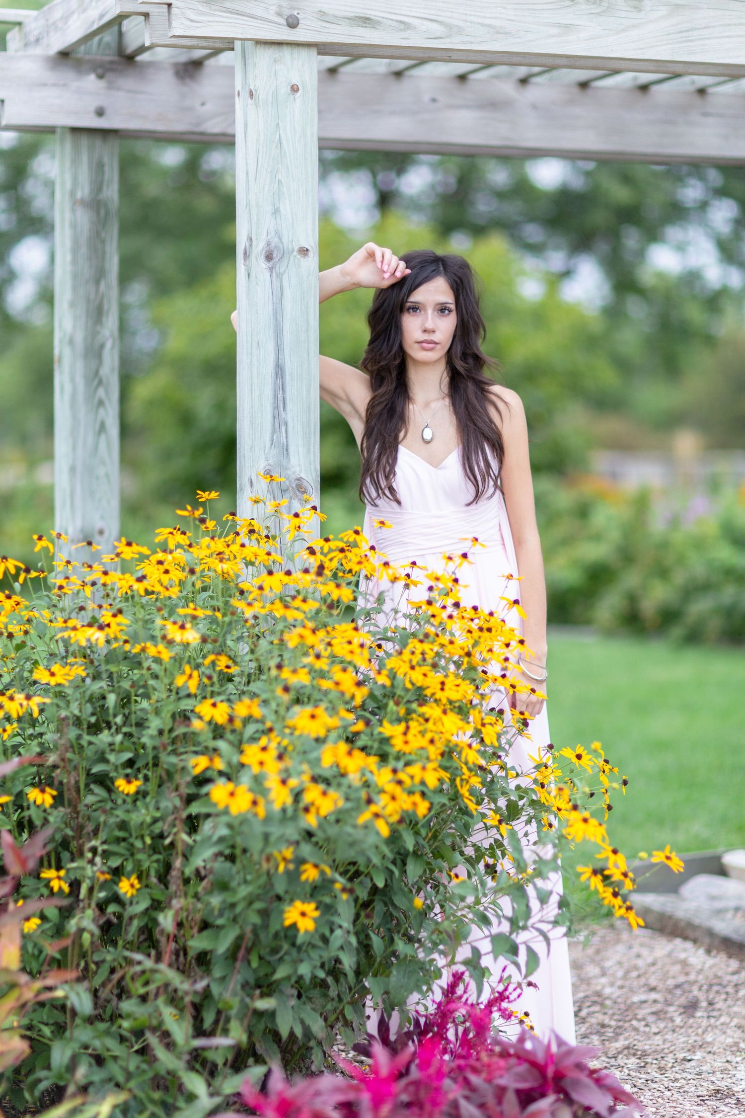 Young woman in white maxi dress posing near wooden pergola surrounded by yellow black-eyed Susan flowers in garden.