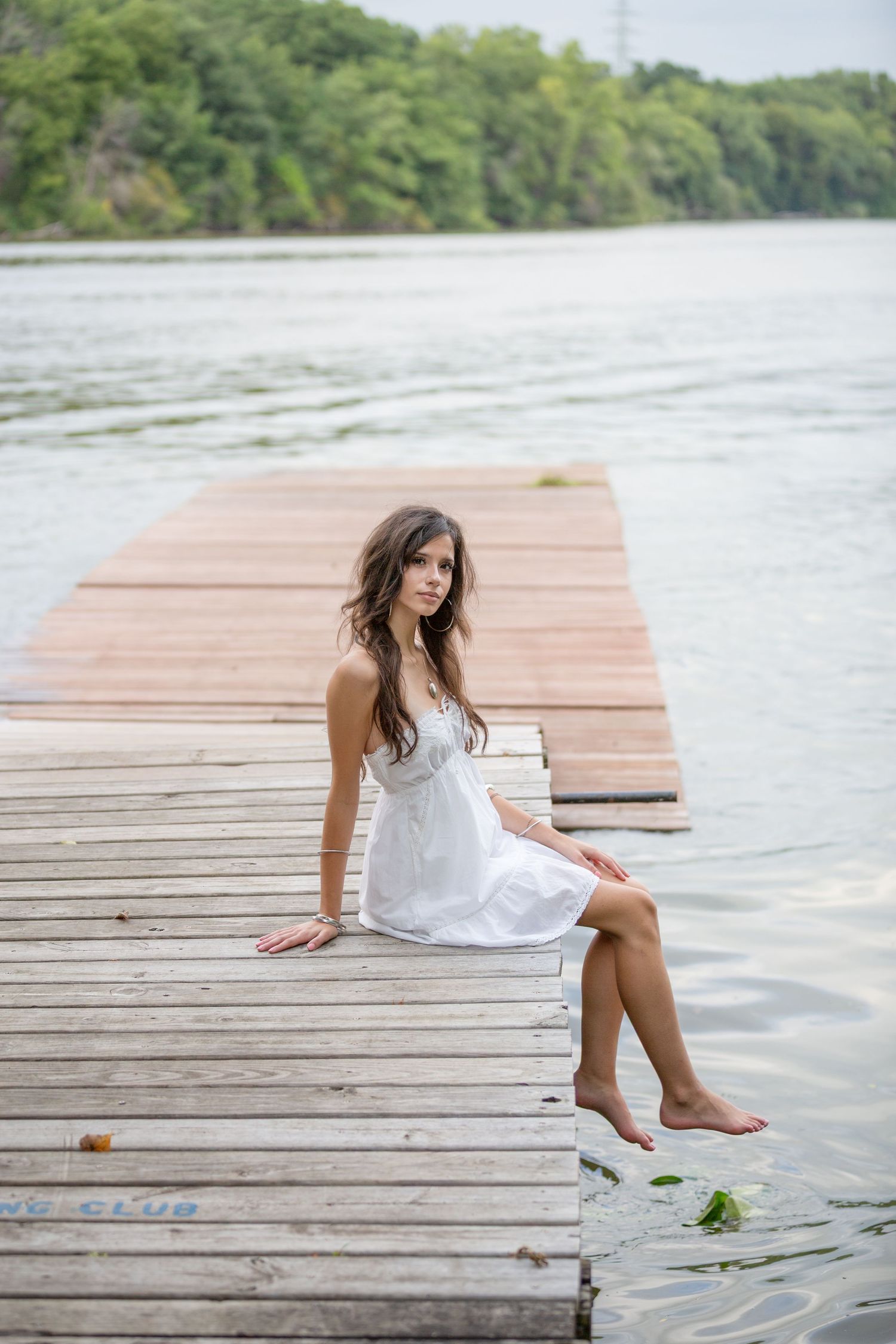 Young woman in white dress sitting barefoot on wooden dock by calm lake with lush green trees in background.