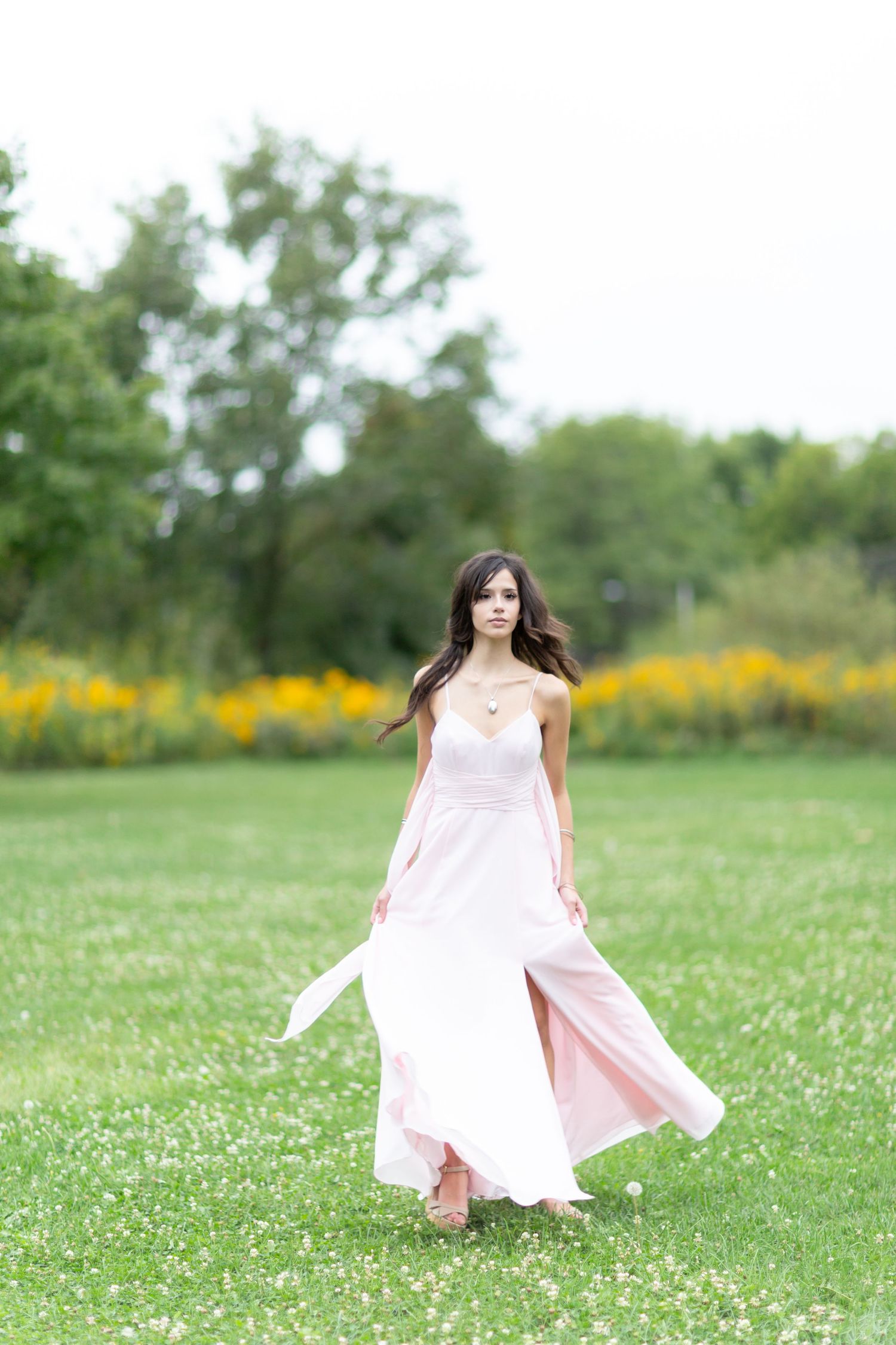 Young woman in a flowing pink dress walking gracefully through a lush green park with yellow flowers in the background.