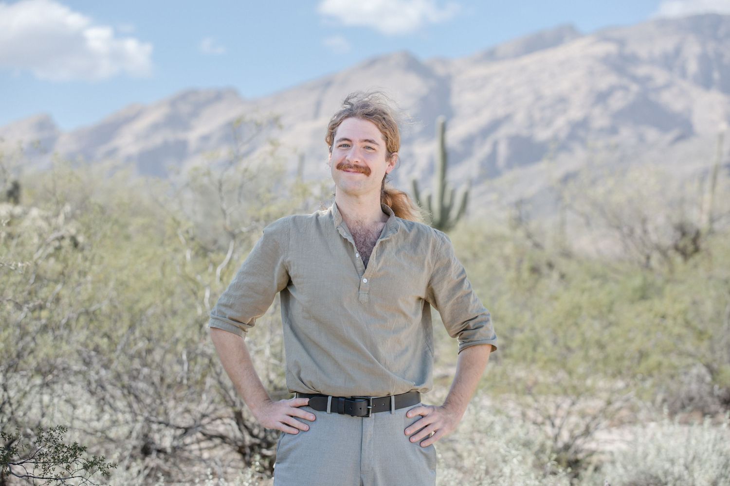 Young man with mustache in olive shirt standing in desert landscape with mountains behind.