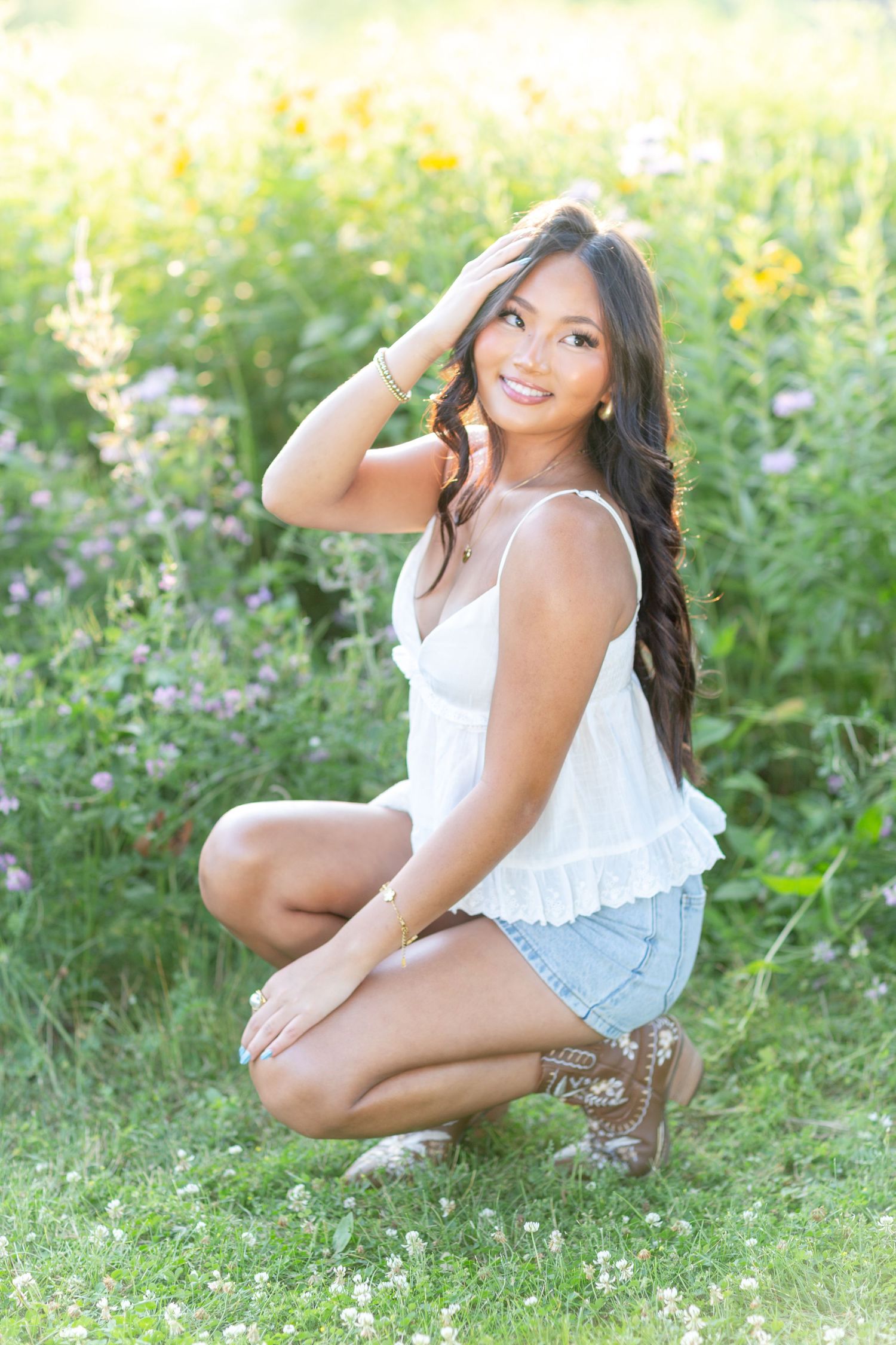 Young woman in white top and denim shorts crouching in a wildflower meadow, smiling over her shoulder.