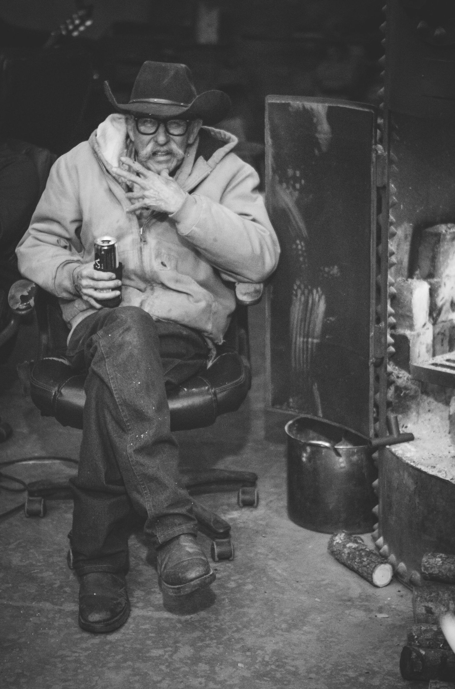 Elderly man in cowboy hat and glasses sitting in workshop chair holding beer can in black and white photo.