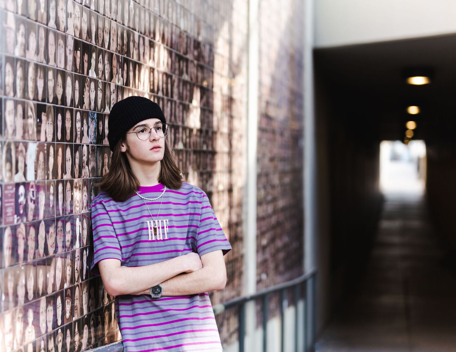 Teenager in striped shirt and black beanie leaning against brick wall in urban corridor.