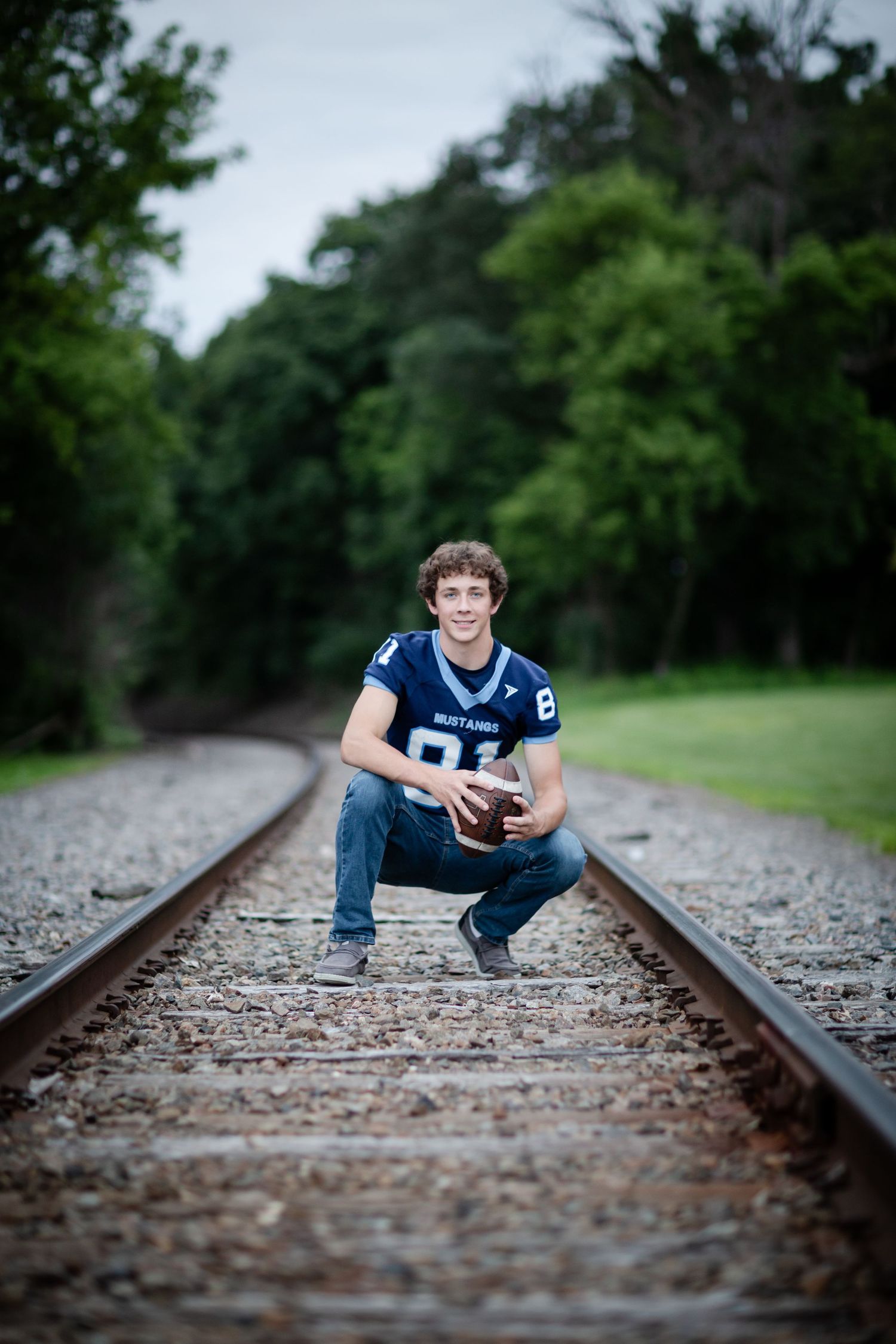 Teen boy in Mustangs football jersey crouching on railroad tracks holding a football, surrounded by green trees.