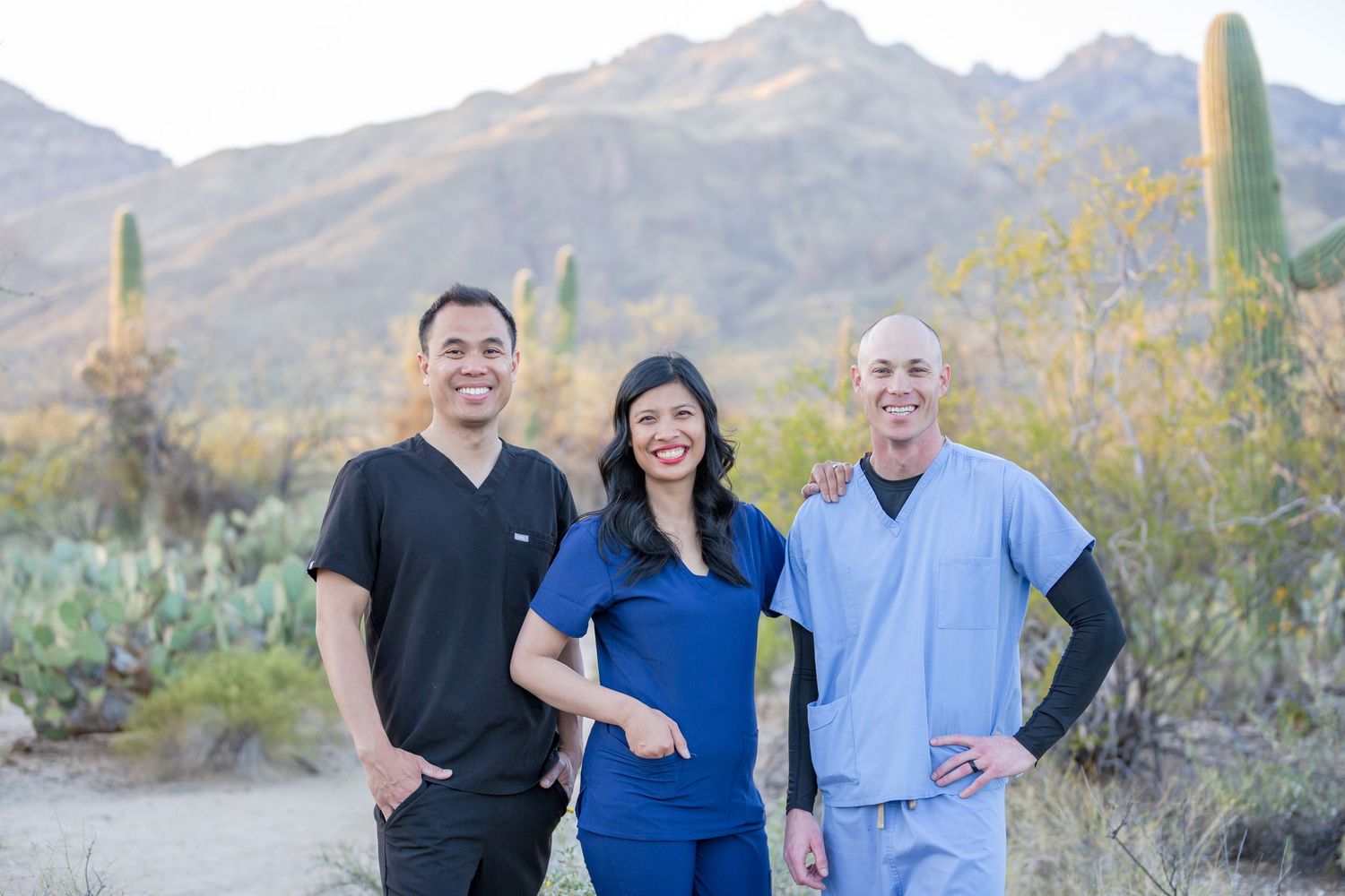 Three medical professionals in scrubs smiling outdoors with desert mountains and cacti in background.
