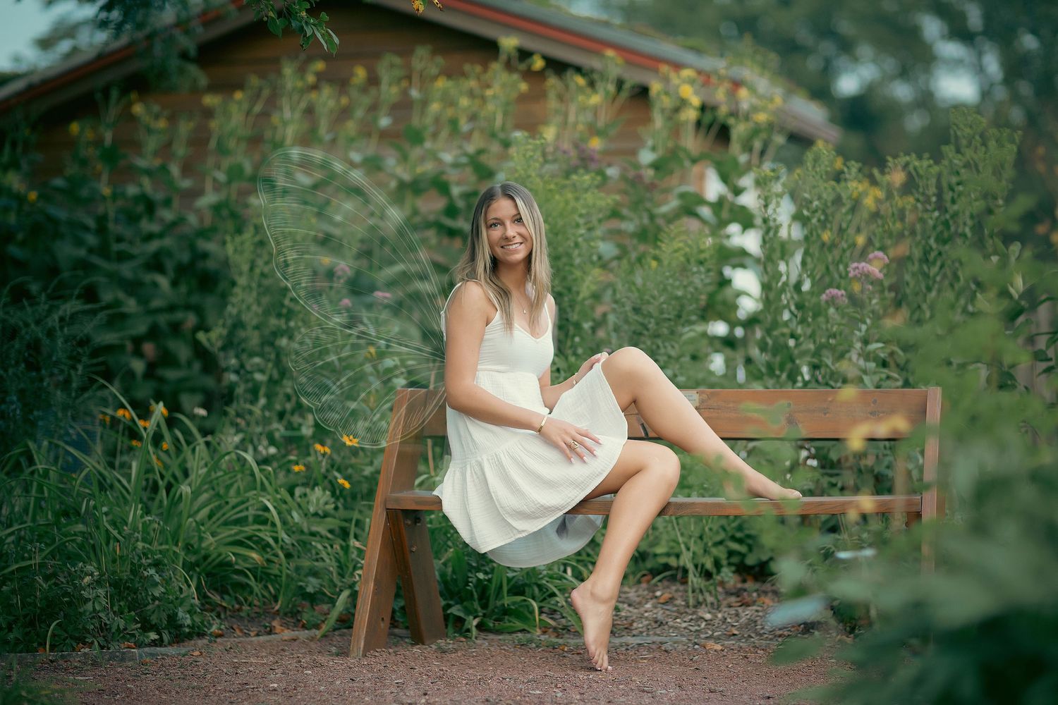 Woman in white dress sitting barefoot on wooden bench surrounded by lush garden greenery near rustic building.