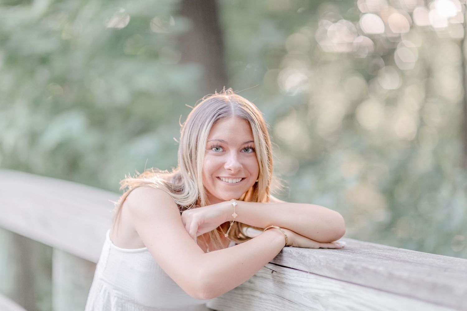 Smiling young woman with blonde hair leaning on a wooden railing outdoors in soft natural light.