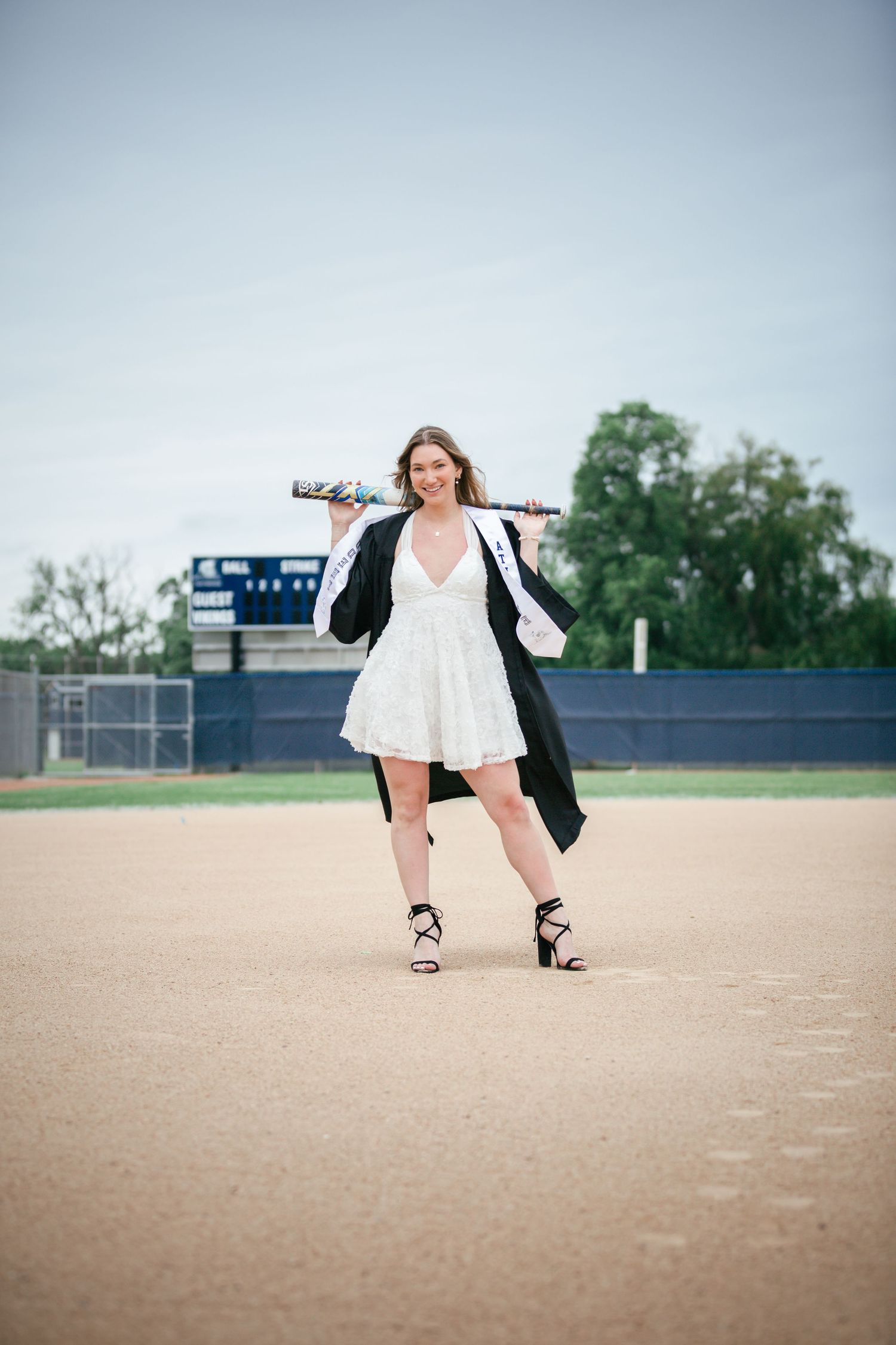 Young woman in white dress and graduation gown holds baseball bat on a dirt field, posing confidently in heels.