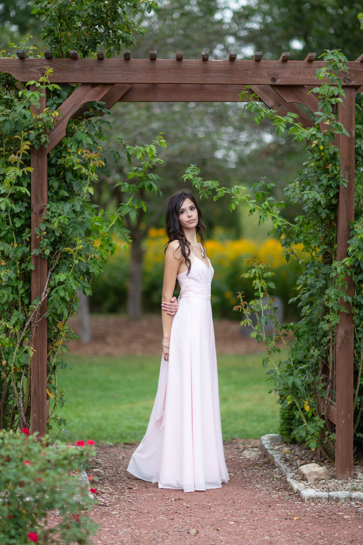 Elegant woman in pink dress posing under wooden pergola arch in lush garden setting.