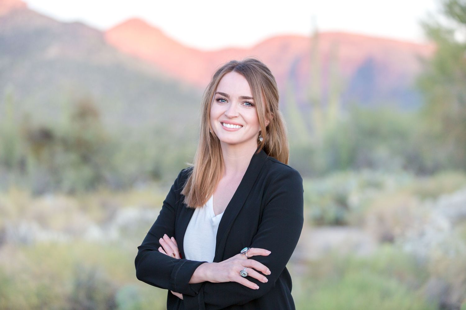 Professional woman in black blazer smiling with arms crossed, standing outdoors with desert mountains at sunset.