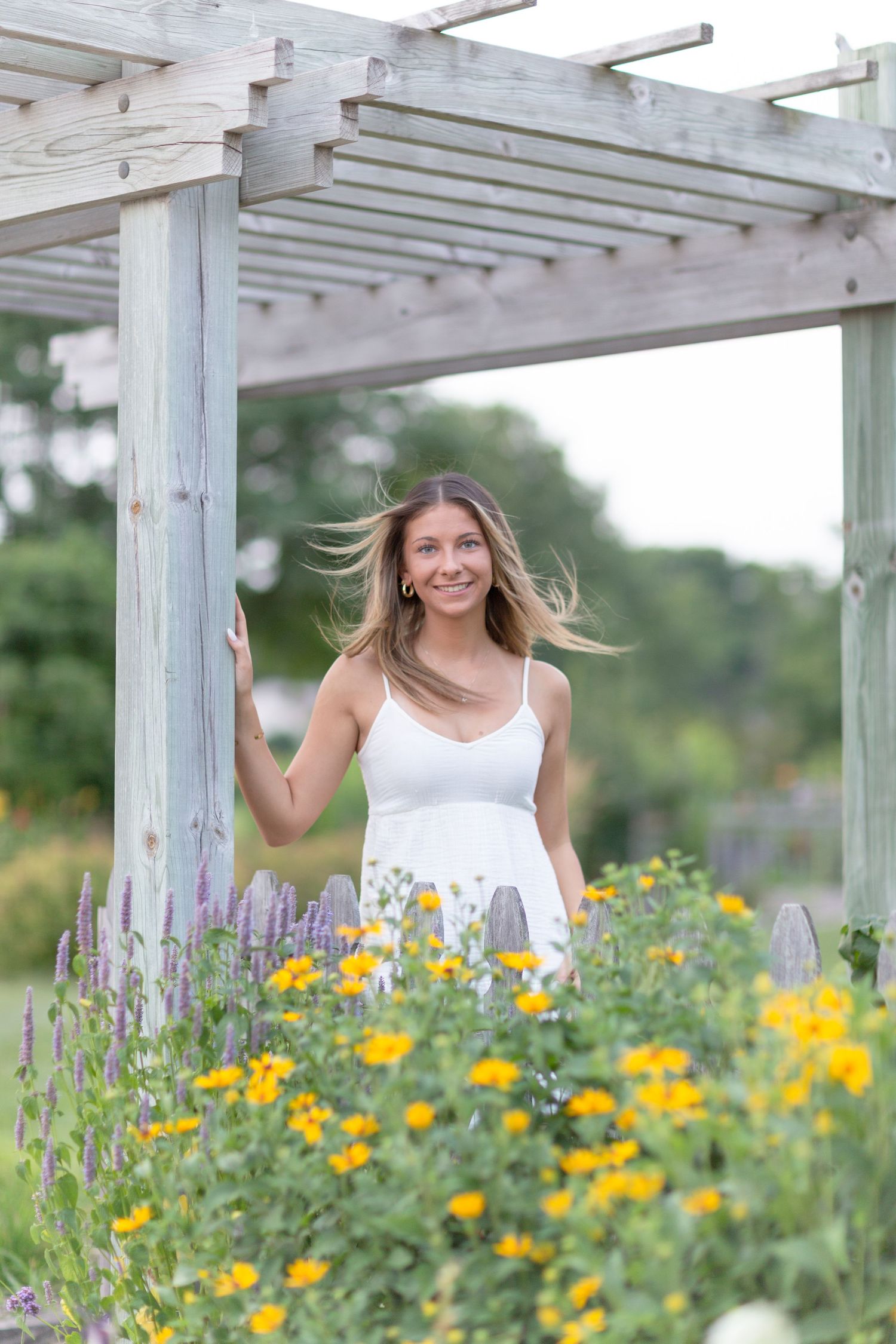 Young woman in white dress smiling among yellow and purple flowers under a wooden pergola.