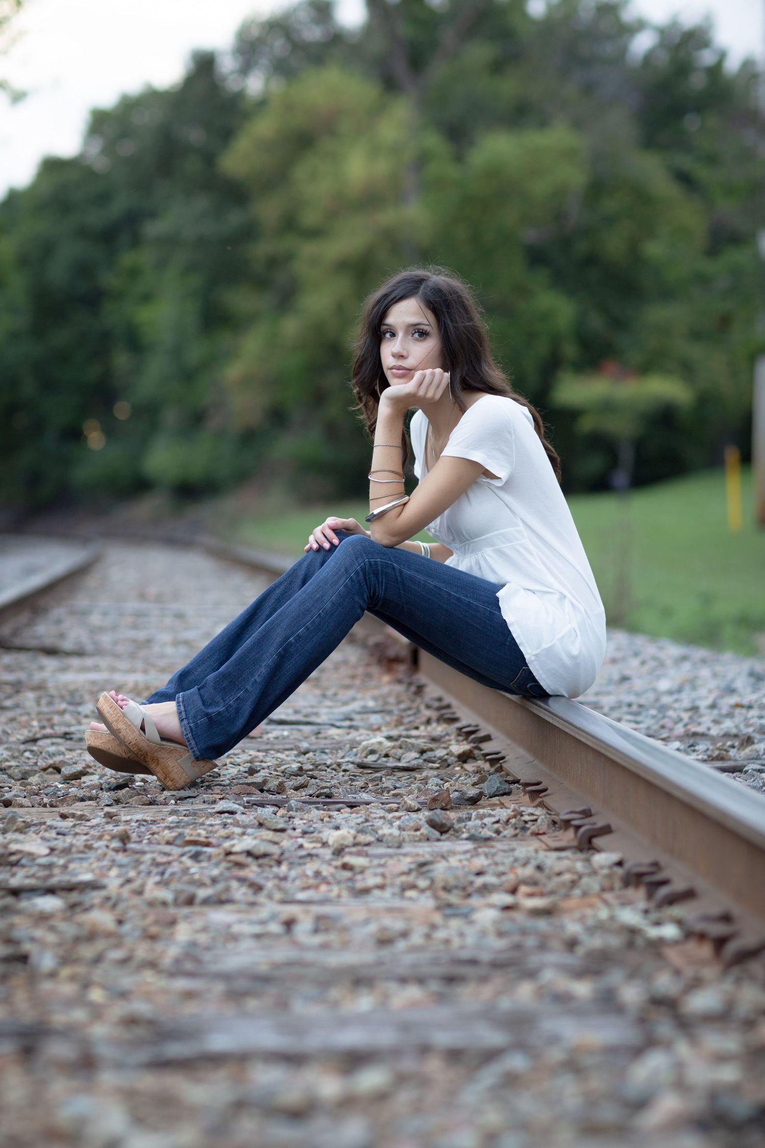 Young woman in white top and jeans sitting pensively on railroad tracks surrounded by lush green trees.