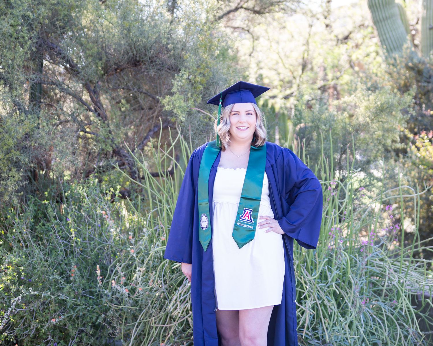 Smiling graduate in blue cap and gown with green stole posing outdoors among desert shrubs.
