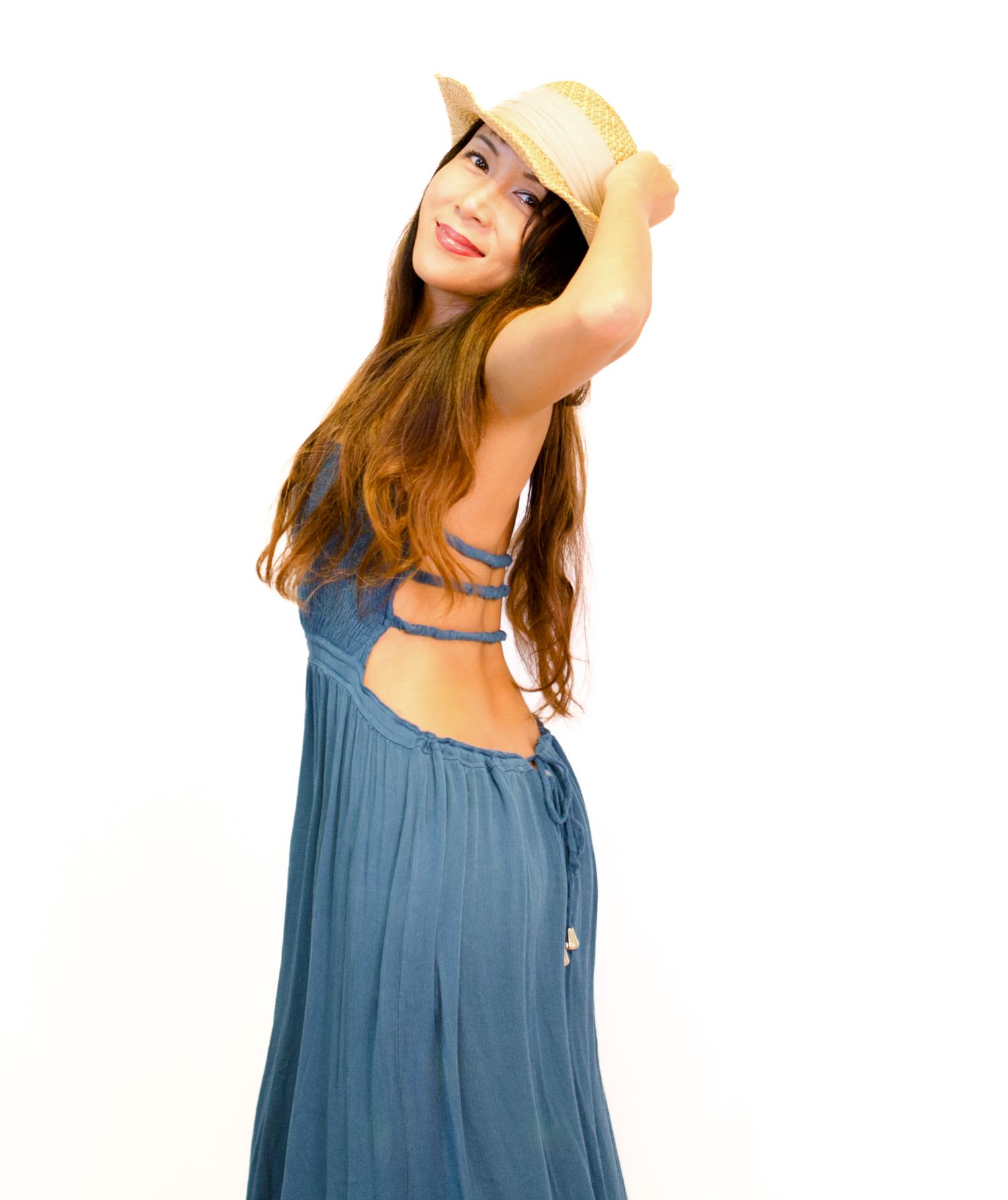 Young woman in a blue backless dress holds a straw hat while posing on a white background.