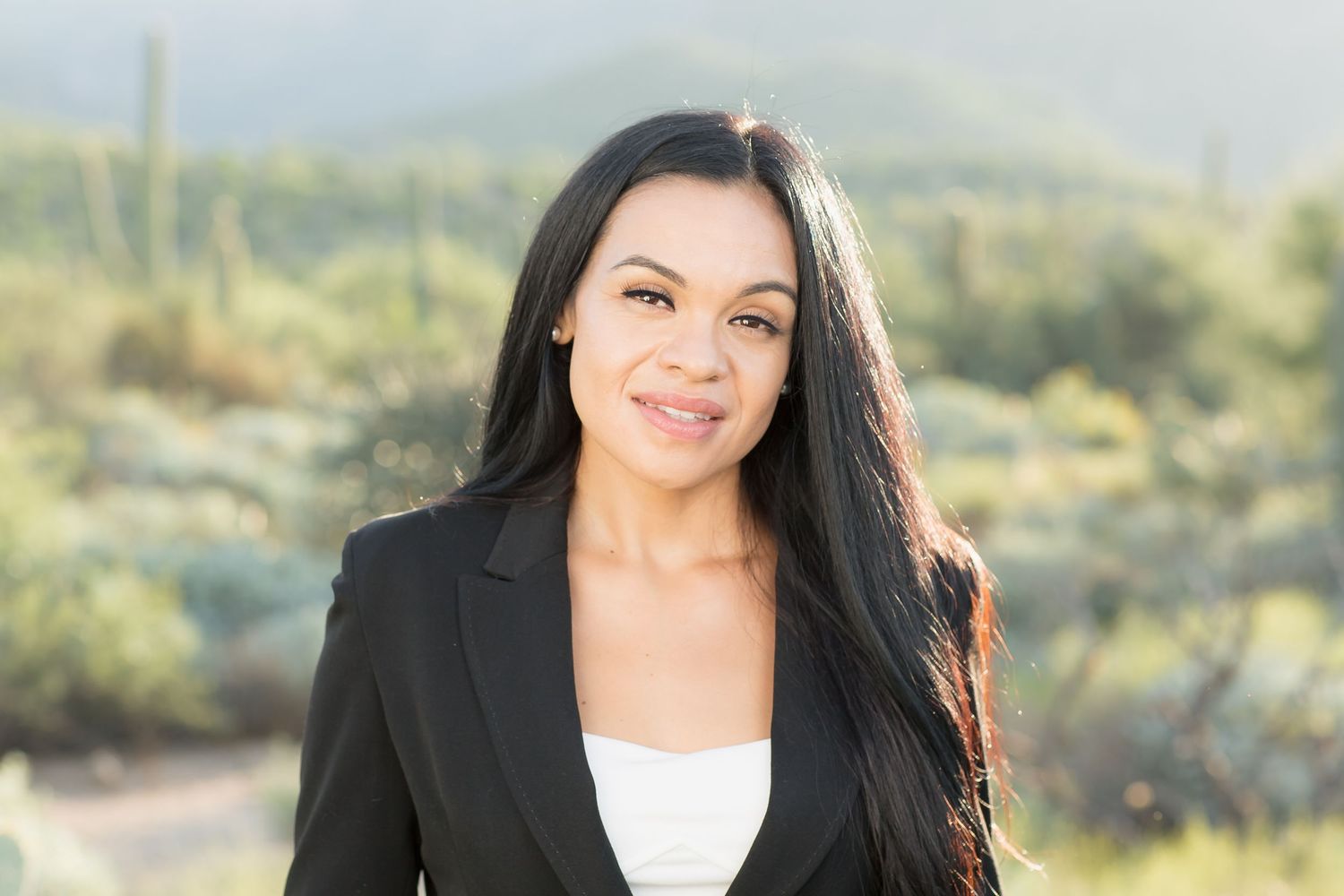 Professional woman in black blazer smiling outdoors with desert cacti and mountains in background.