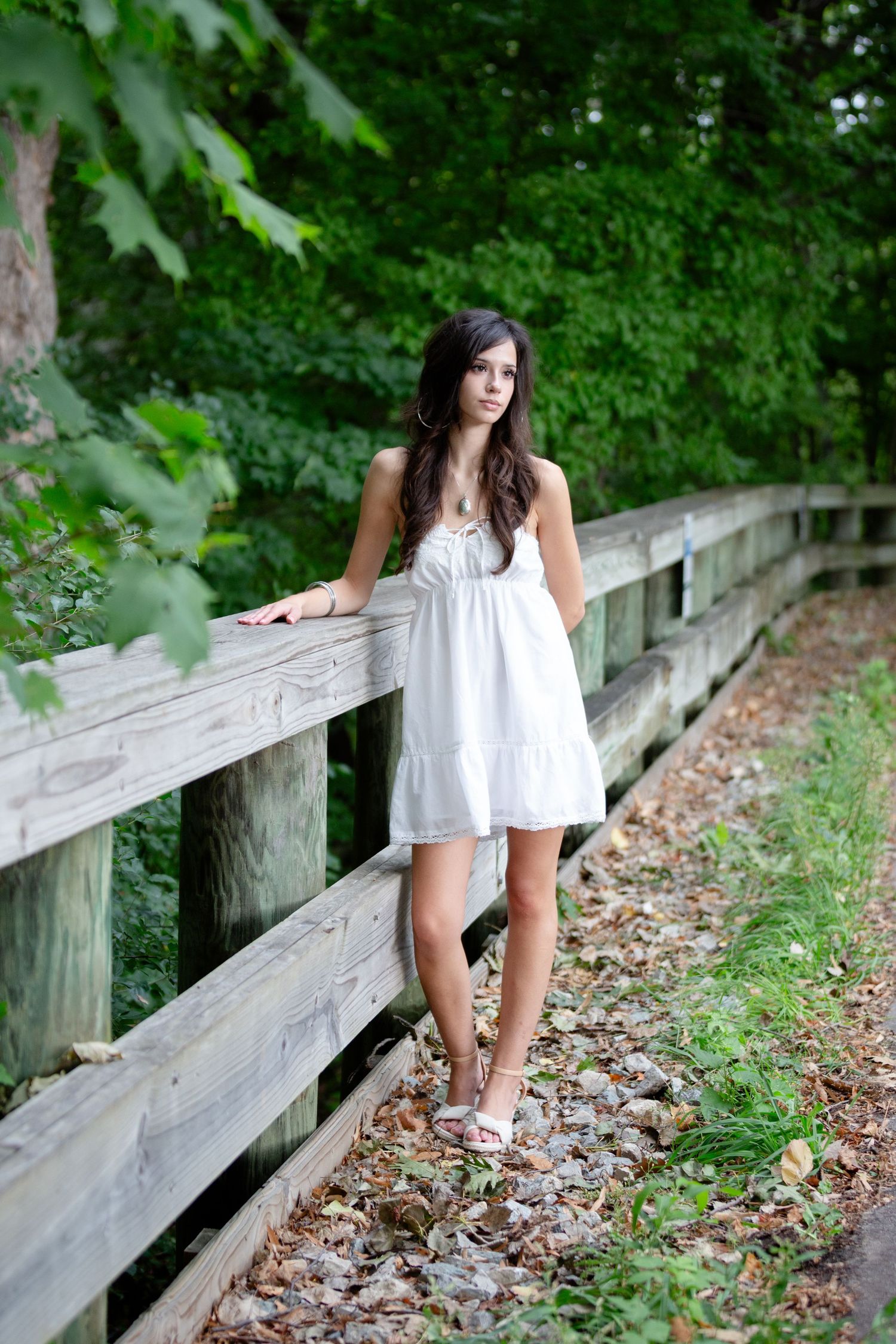 Young woman in white sundress leaning against rustic wooden fence surrounded by lush green trees and fallen leaves.