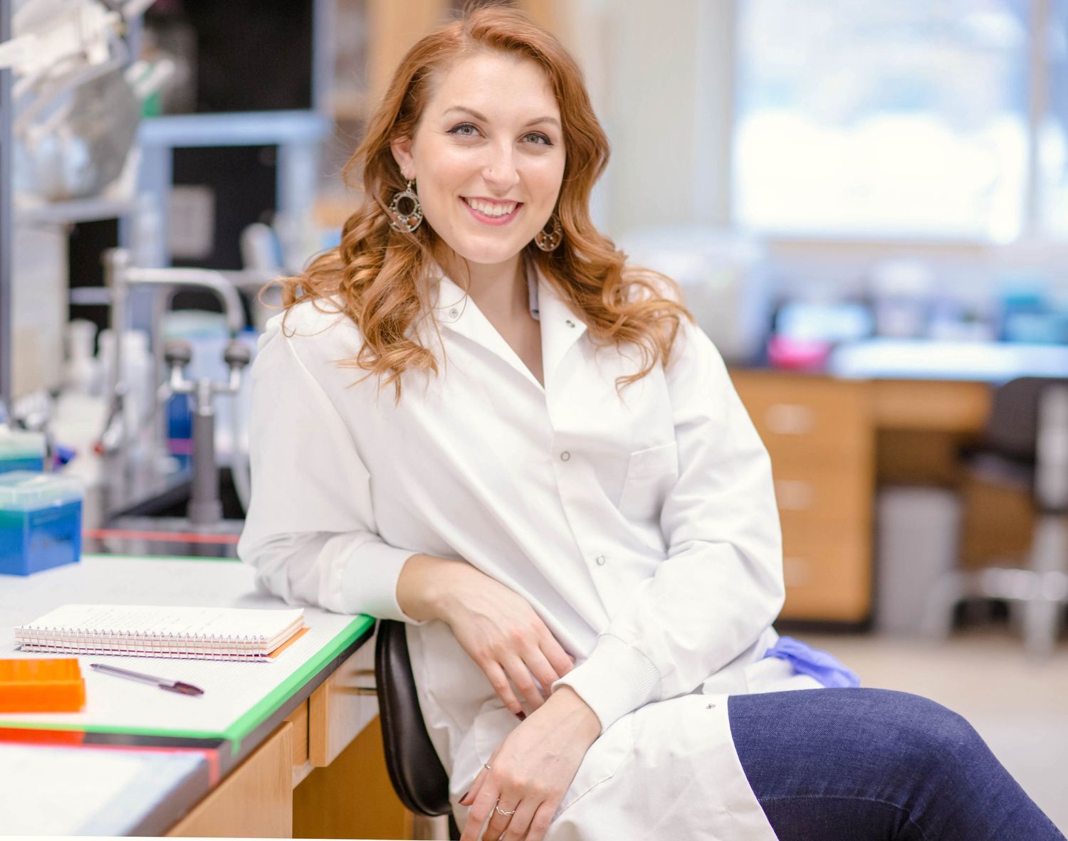 Smiling female researcher in white lab coat seated at desk in bright modern laboratory.