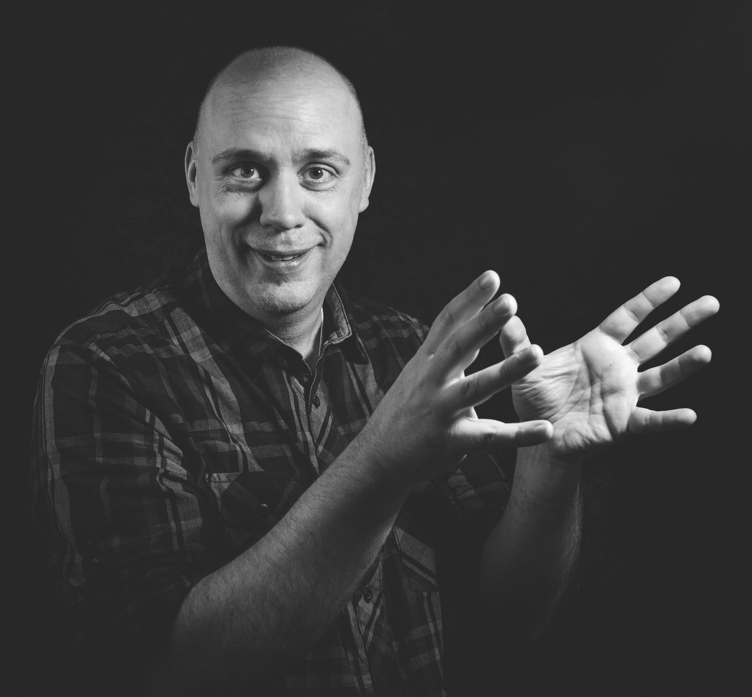 Bald man in plaid shirt gesturing with open hands against dark background in black and white photo.