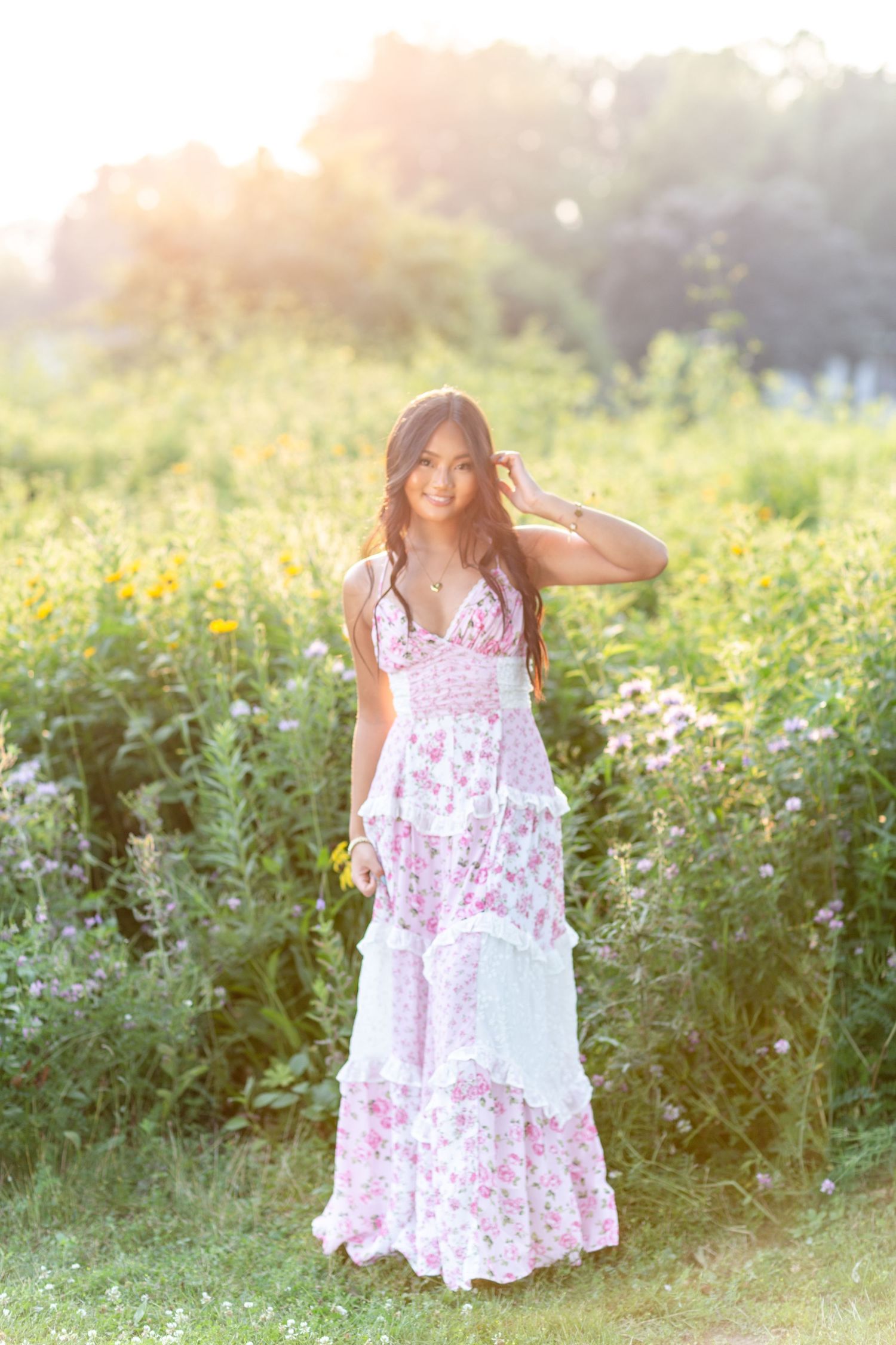 Young woman in floral maxi dress standing in a sunlit wildflower field during golden hour.