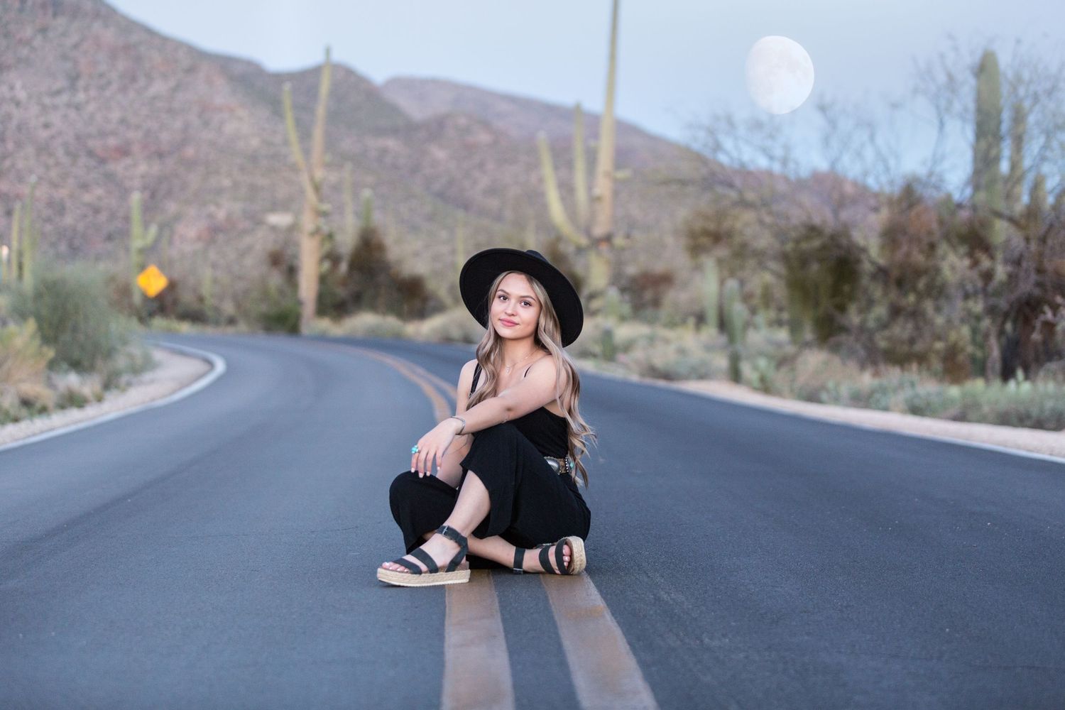 Young woman in black hat and outfit sitting on desert road with saguaro cacti and full moon backdrop.
