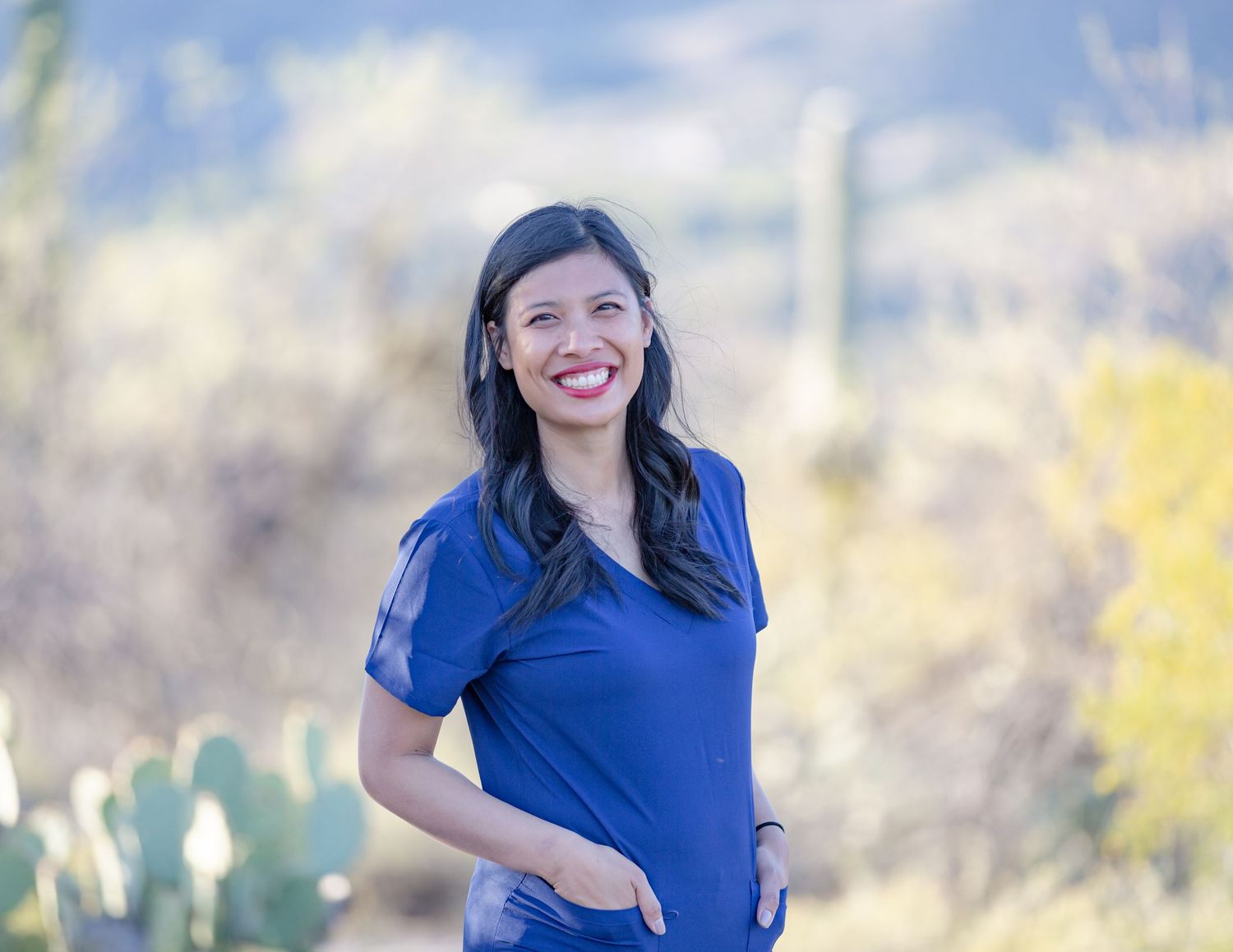Smiling woman in blue scrubs standing outdoors in sunny desert landscape with cacti in background.