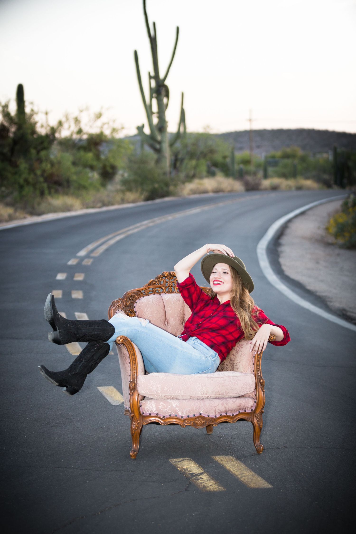 Young woman in red plaid shirt and hat relaxing in vintage chair on open desert road.