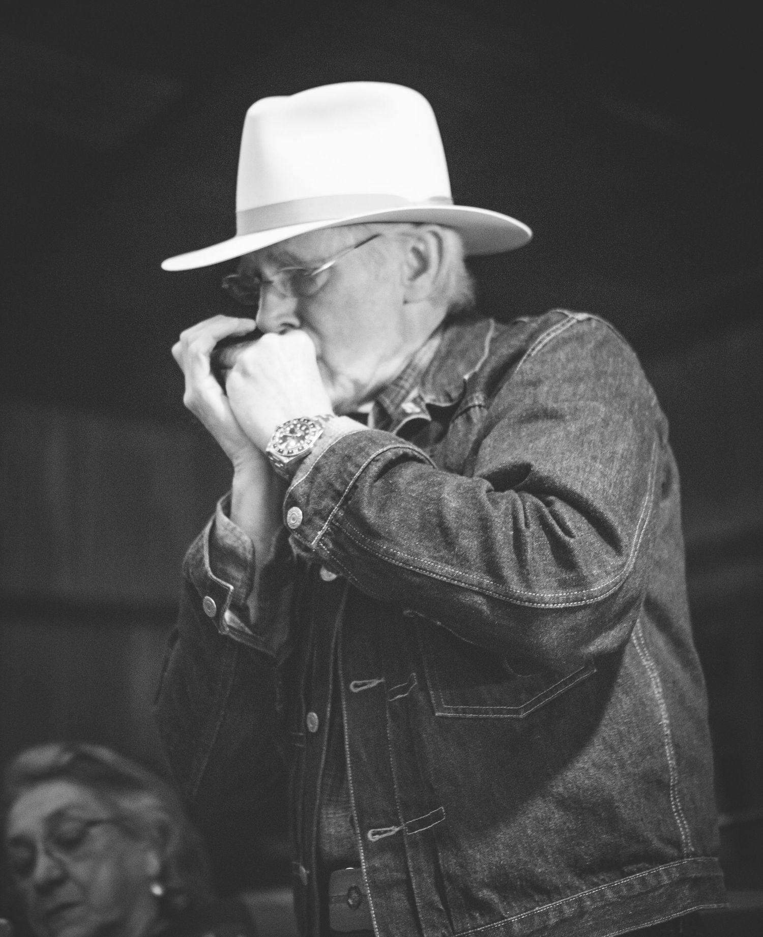 Elderly man in white cowboy hat playing harmonica outdoors in black and white portrait photo.
