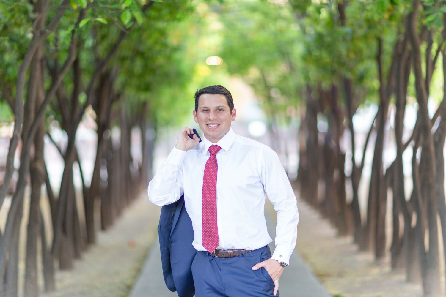 Smiling businessman in white shirt and red tie walking along a tree-lined path.