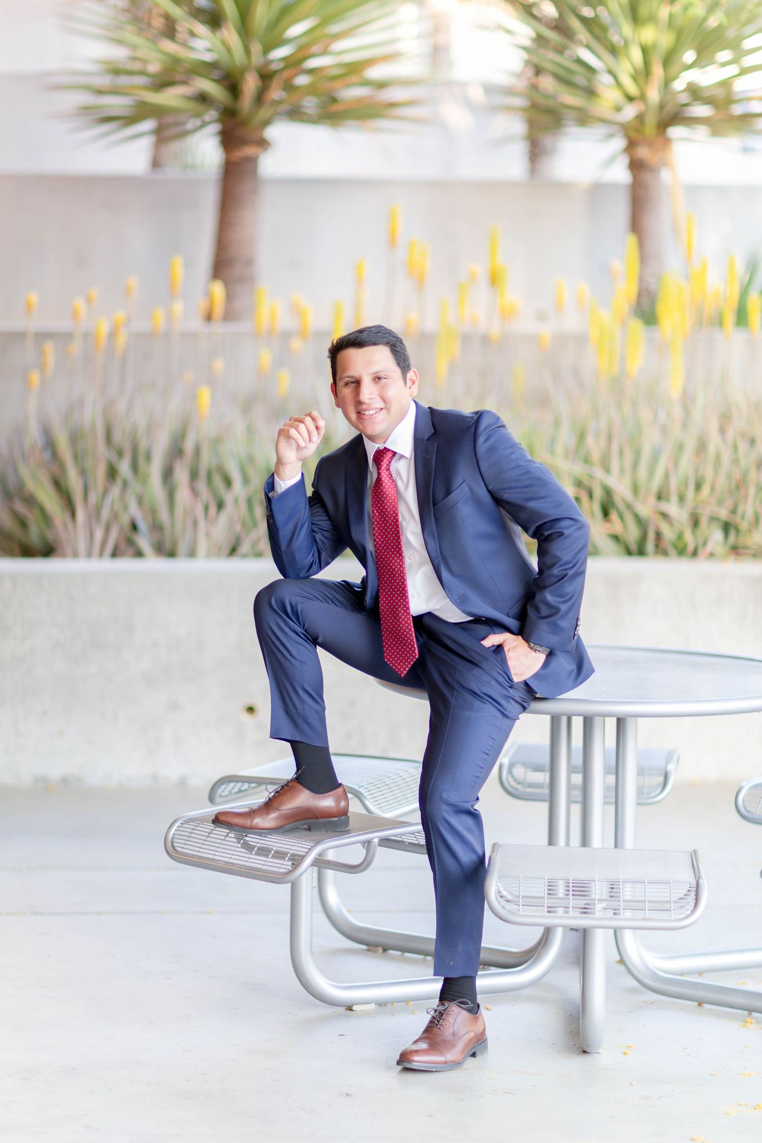 Professional man in navy blue suit and red tie posing at an outdoor table.