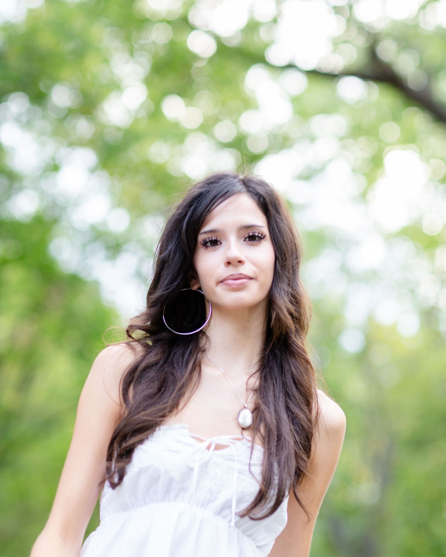 Young woman with long dark hair wearing a white dress and hoop earrings, posing outdoors with green trees.
