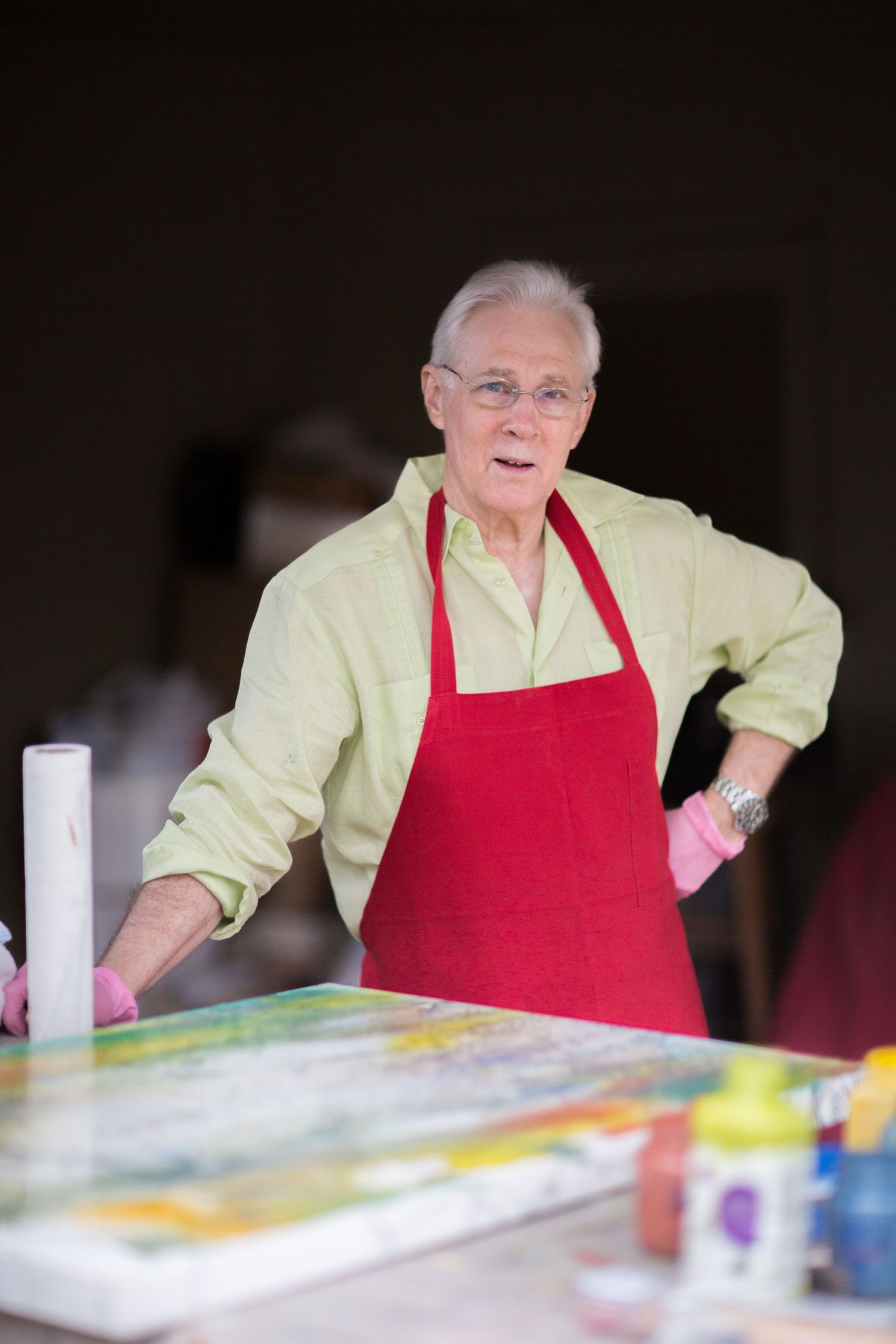 Senior man in red apron and green shirt working on colorful painting at art table.