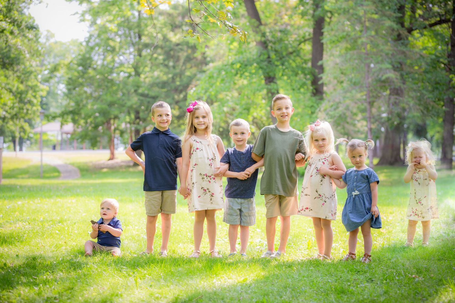 Group of young children posing together on green grass in a sunny park surrounded by trees.